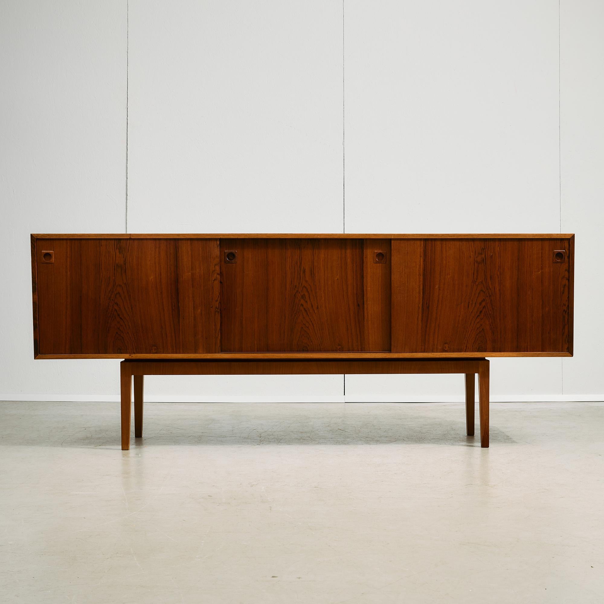 Questa credenza abbina bei materiali a un design accurato. Il corpo è realizzato in impiallacciatura di palissandro riccamente figurato, su un telaio sottile e rialzato in teak massiccio che mantiene la silhouette leggera e raffinata. Tre ante