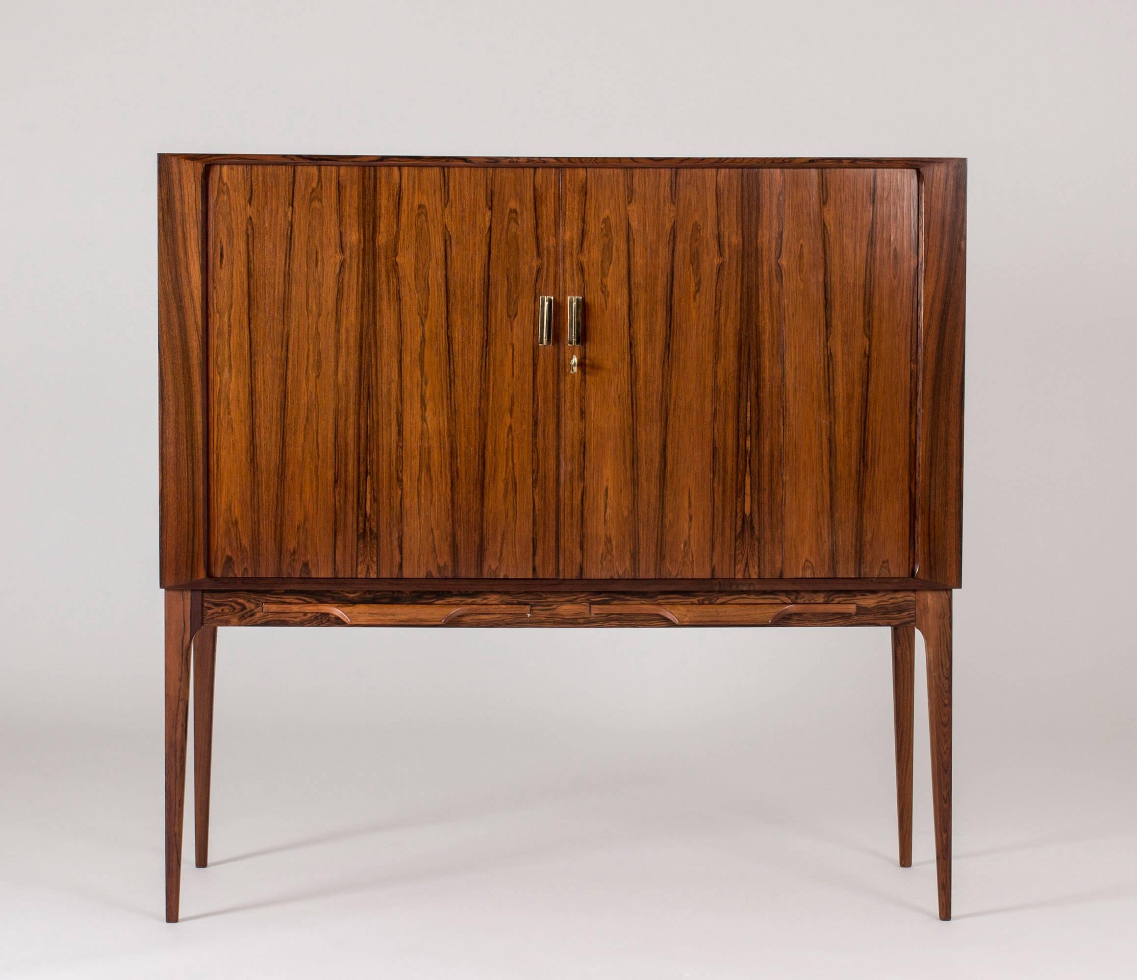 Beautiful rosewood bar cabinet by Kurt Østervig with jalousie doors and a dramatic veneer pattern. Two bakelite dressed trays can be pulled out underneath the cabinet doors. Mirrored inside with a long glass shelf on one side and white lacquered
