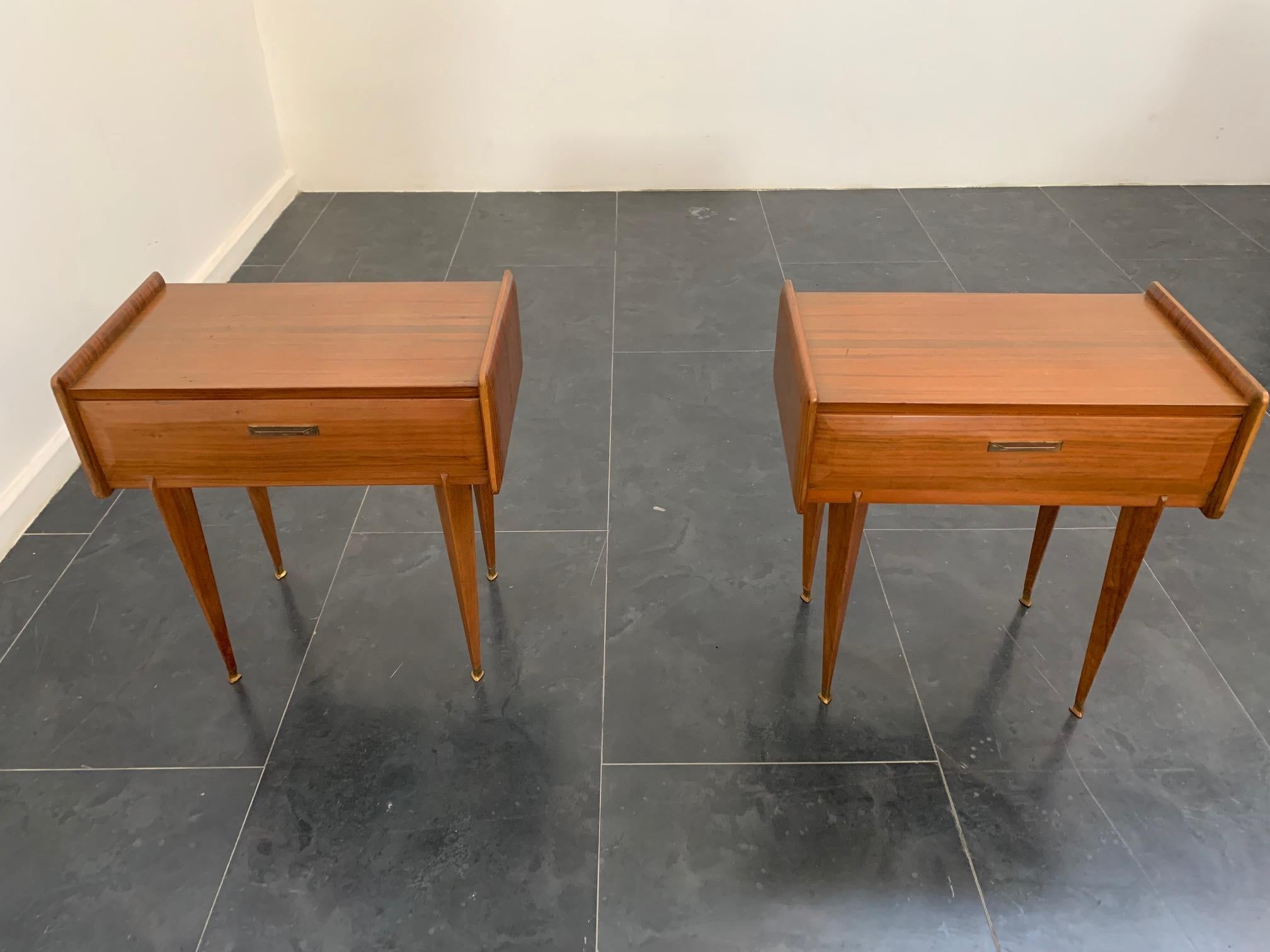 Pair of rosewood bedside tables, 1950s. Featuring pyramidal spiked legs with elegant brass tips. The sides are shaped and domed and extend to accommodate the body and top. The front features a rusticated drawer with a gilded metal handle. The wood