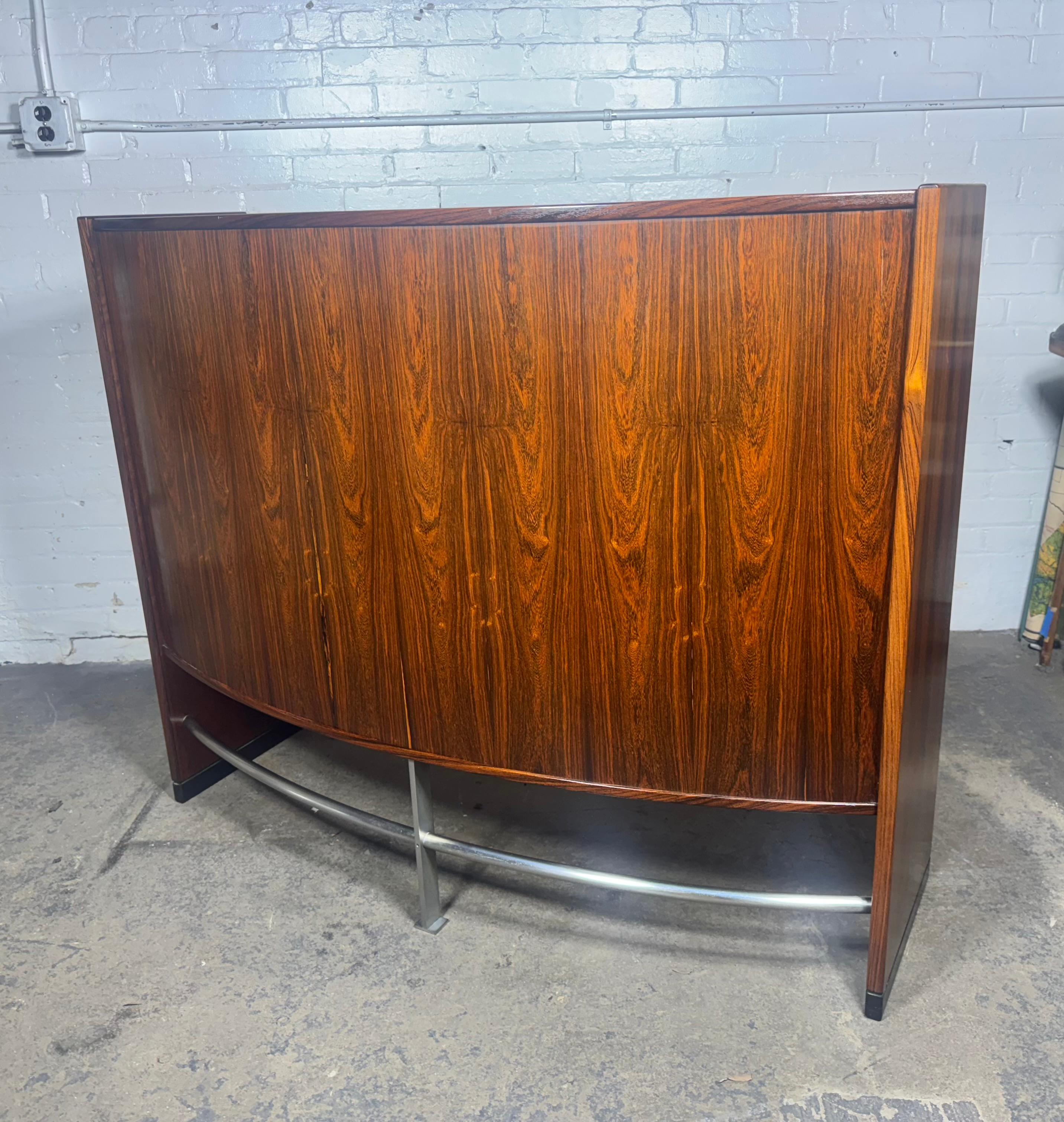 This Danish Modernist boomerang dry bar by Erik Buch for Dyrlund is a stunning piece from the 1960s. Seldom seen in rosewood.. Its striking boomerang-shaped front and chrome-plated steel footrest exude mid-century modern charm. The back features a
