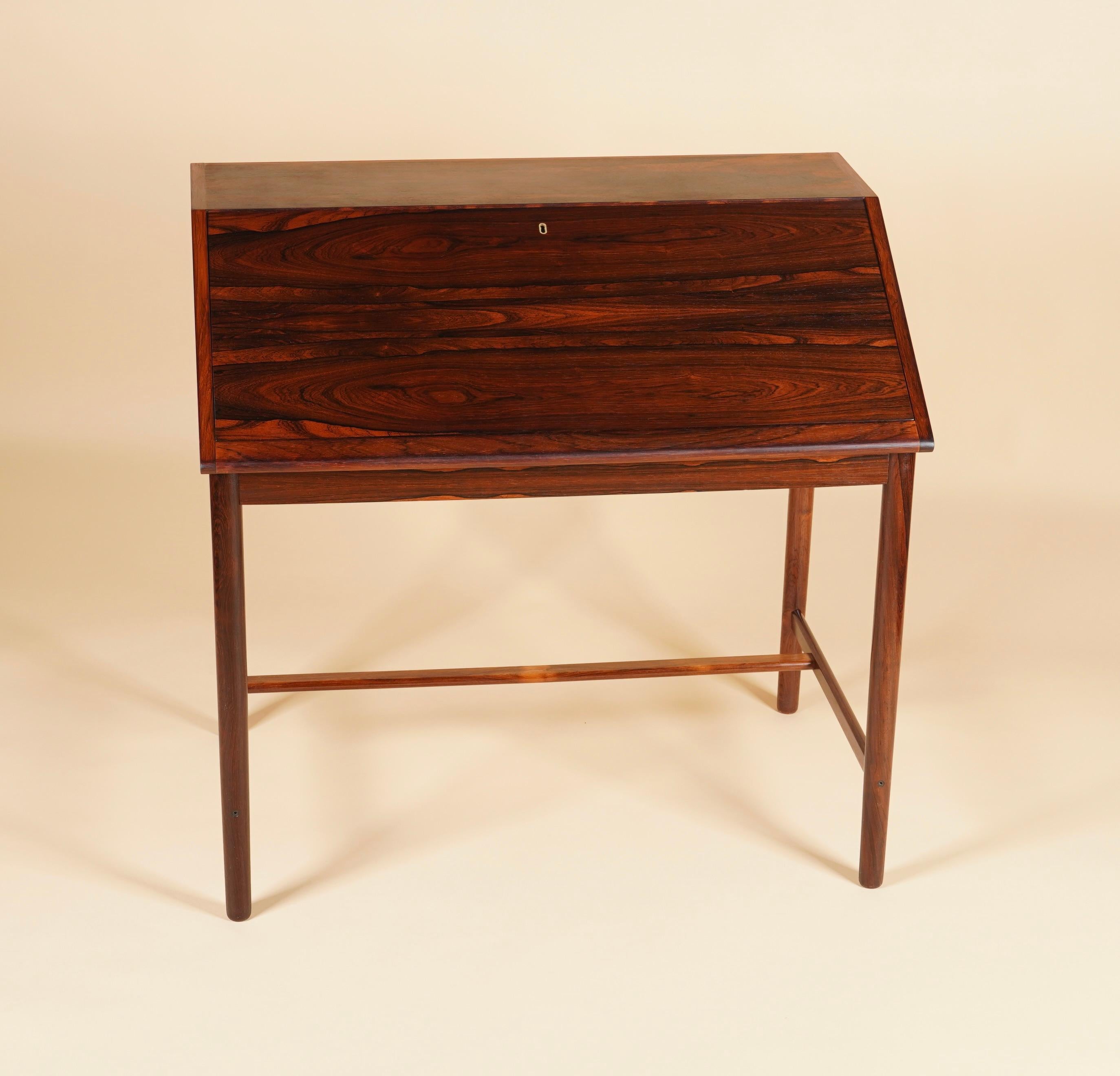 Compact drop front desk by Brode Blindheim of Norway, with an angled profile and clean architectural lines of the era. Crafted in rosewood with rich reds and striking black grain. Inside are two small drawers and cubbies for paperwork storage. Keys