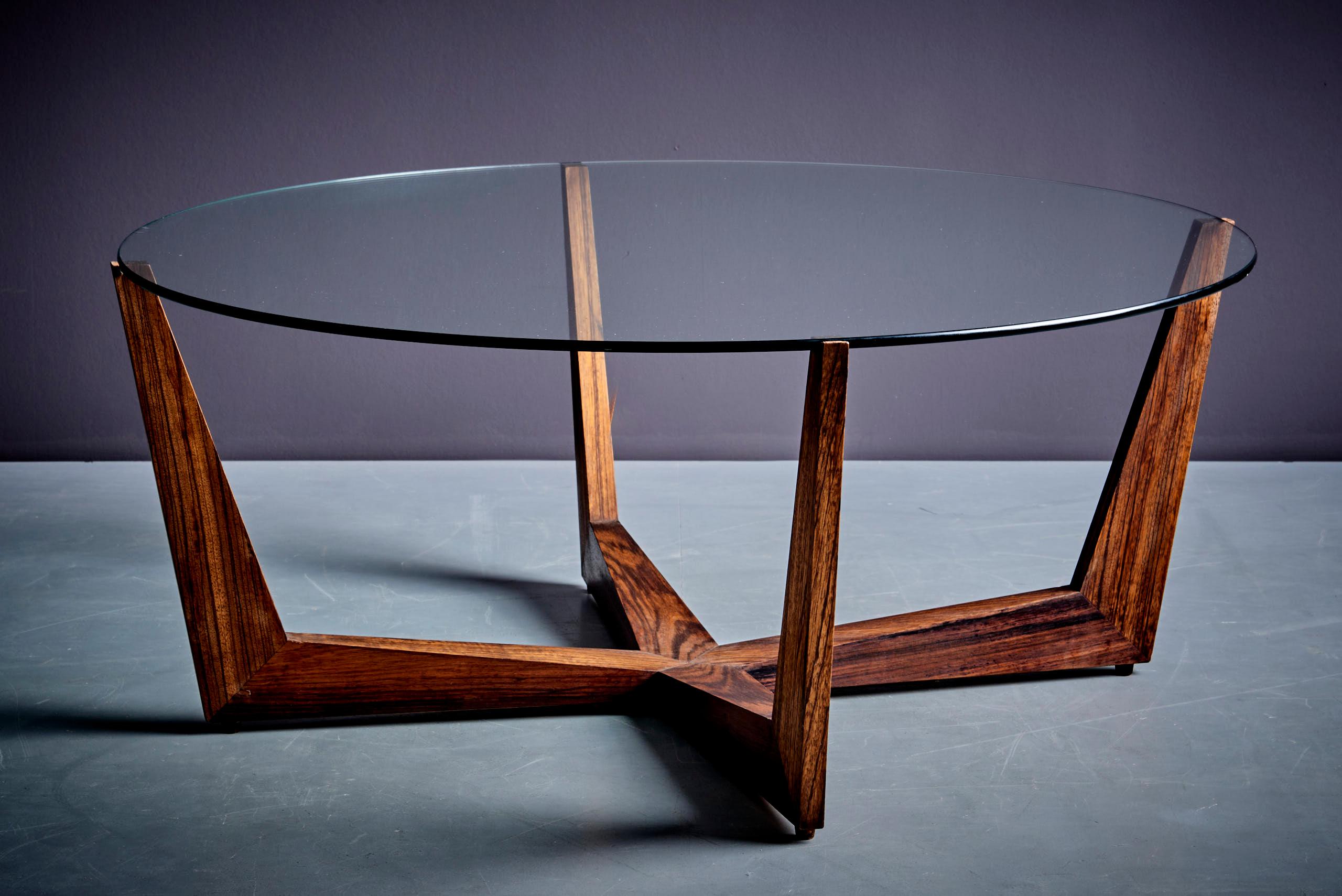 This table, designed by Wilhelm Renz, features a round glass top supported by a sculptural rosewood base. The wooden base is geometric and angular, giving the table stability and a modern look. The glass top contrasts with the warm, natural wood
