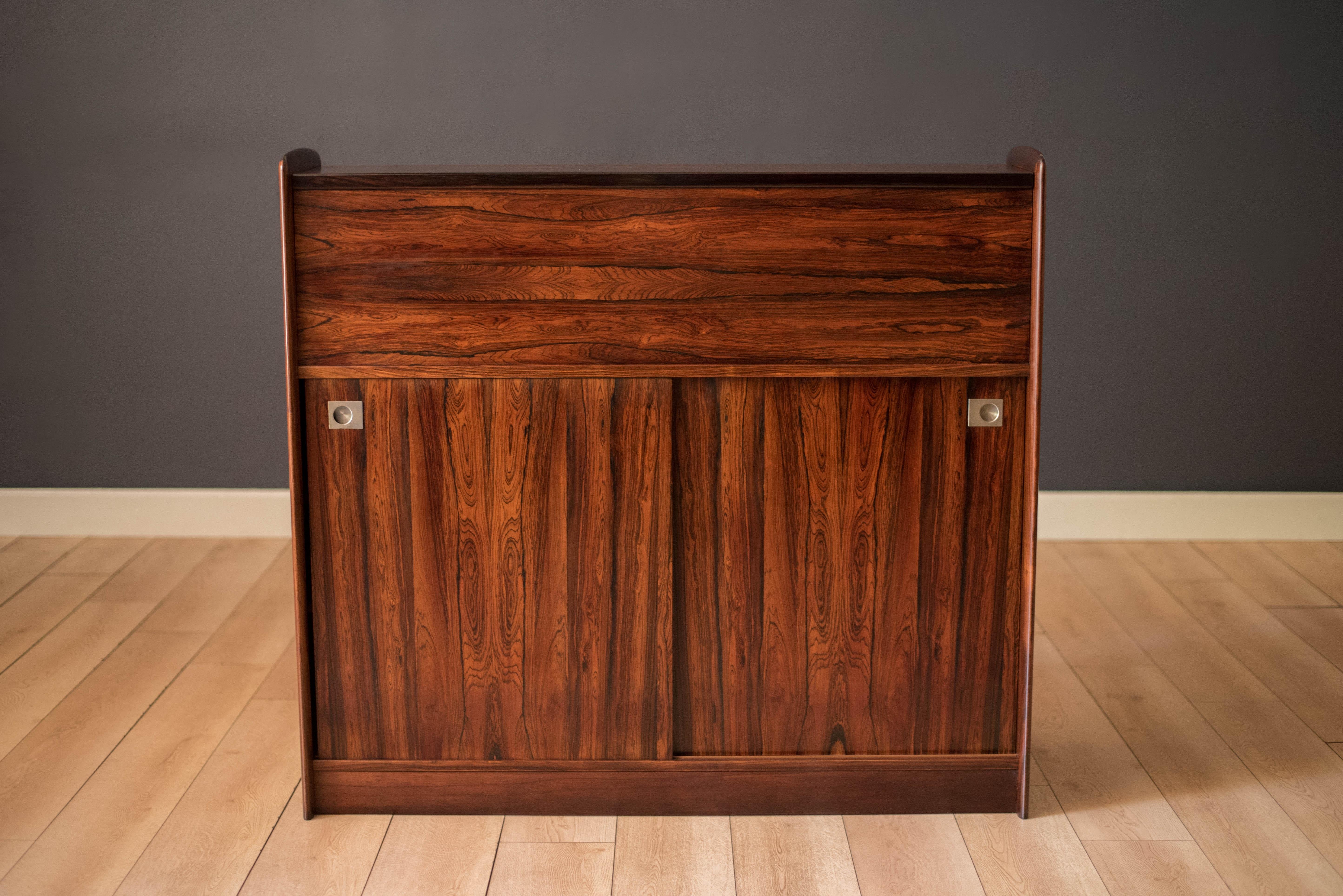 Mid-Century Modern dry bar with sideboard cabinet in rosewood circa 1970’s. Features an expandable serving countertop with an easy to maintain black laminate surface. The interior bar is equipped with one removable shelf, five bottle holders, a