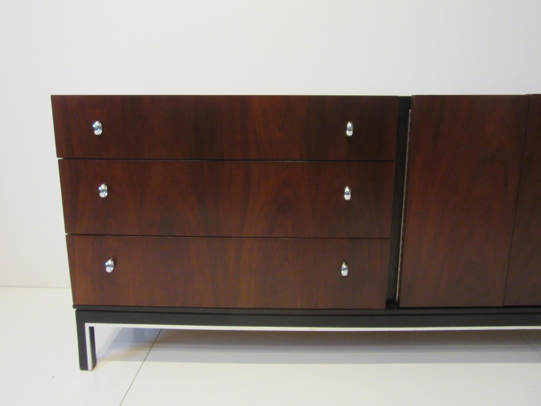 A richly grained rosewood and ebony toned dresser / credenza with tri fold doors revealing three drawers and the other side having three drawers with chrome pulls. The stretcher and leg area is detailed with an aluminum strip giving the piece a very