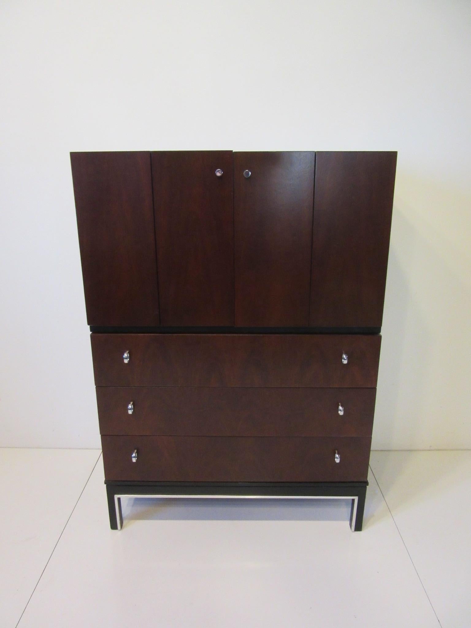 A well grained rosewood and ebony toned tall dresser chest with upper bi fold doors having storage sections and a drawer. The lower area has three drawers with chrome pulls and aluminum trim to the stretcher and leg area, retains the manufactures
