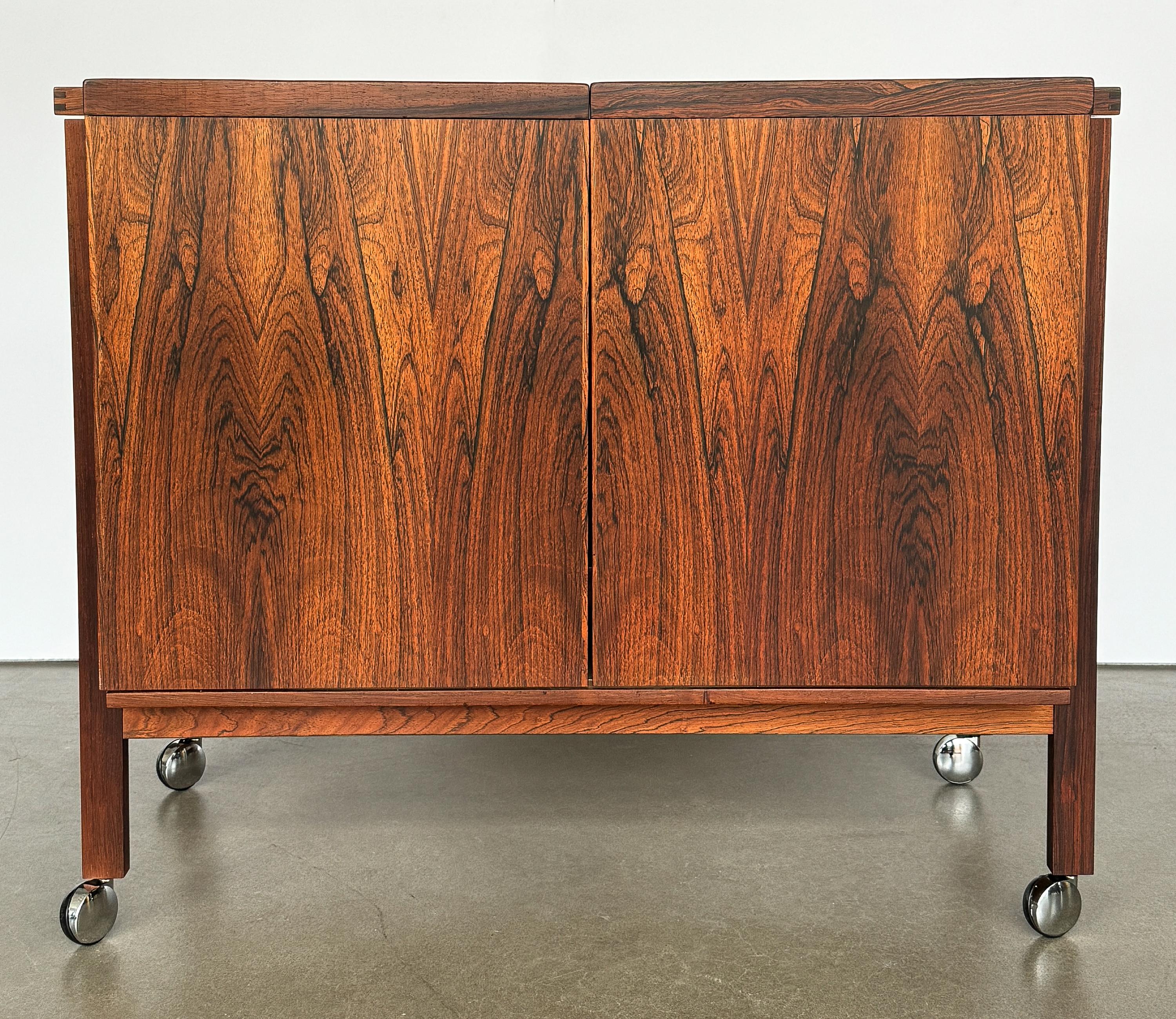 This Danish Mid Century expandable rosewood bar cart / cabinet, designed by Niels Erik and Glasdam Jensen for Vantinge Mobelindustri in the 1960s, is a paragon of Scandinavian design—where functionality meets the pinnacle of style. The beautiful