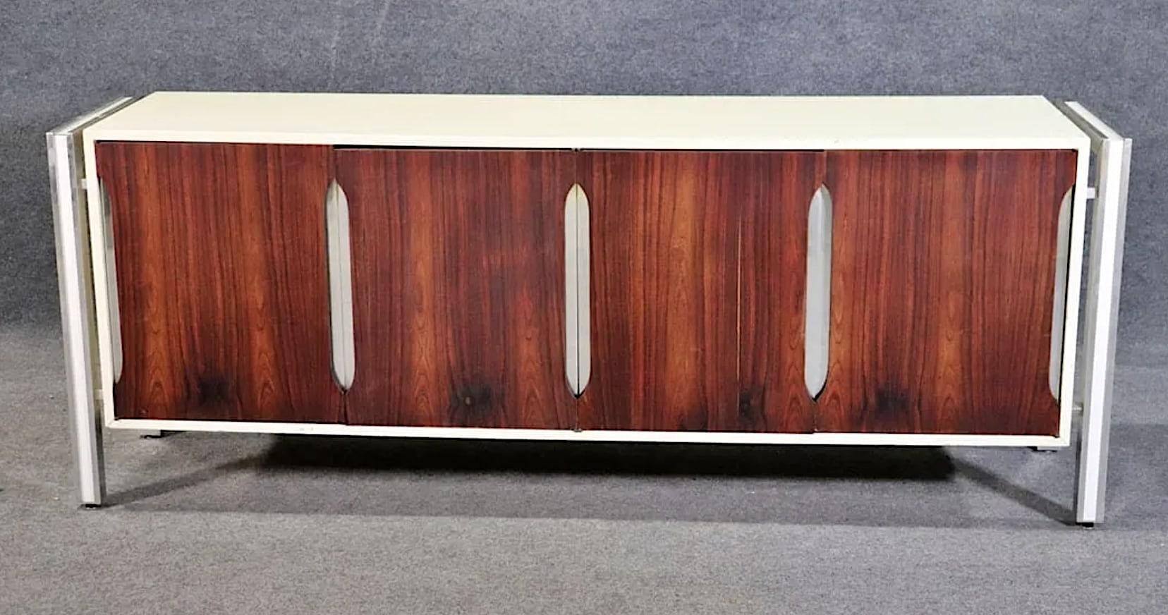 Long mid-century space age dresser with stunning features. Four rosewood lined doors with polished aluminum trim and frame, contrasting white body. Six deep drawers sit behind the rosewood doors. 
Please confirm location NY or NJ 