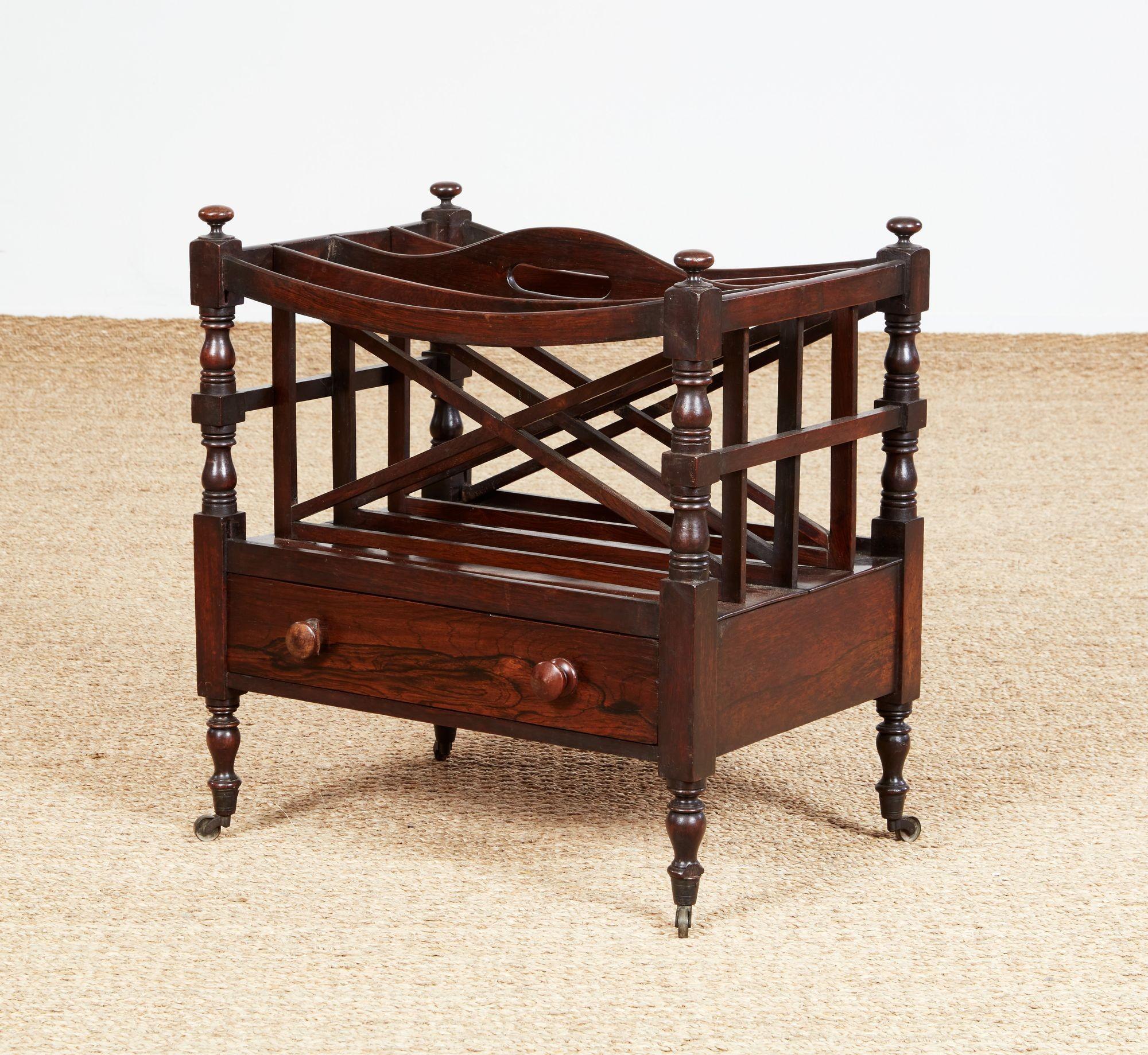 A magazine rack, or canterbury, fashioned of rich rosewood timber, having four filing slots with removable dividers in a fixed frame with a central cutout handle, above a single drawer, standing on turned legs ending in brass casters.