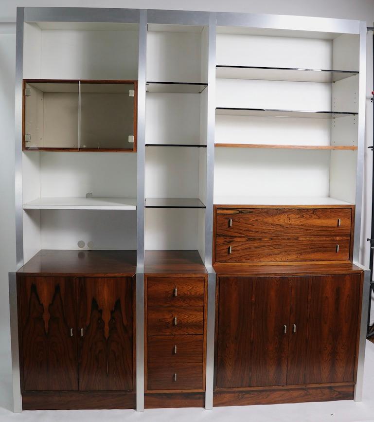 Stunning wall unit having rosewood cabinets with white Formica surfaces, brushed metal trim, and tinted glass shelves. Impressive three bay unit, having storage cabinets, drawers and a glass fronted display case. This example is in very good,