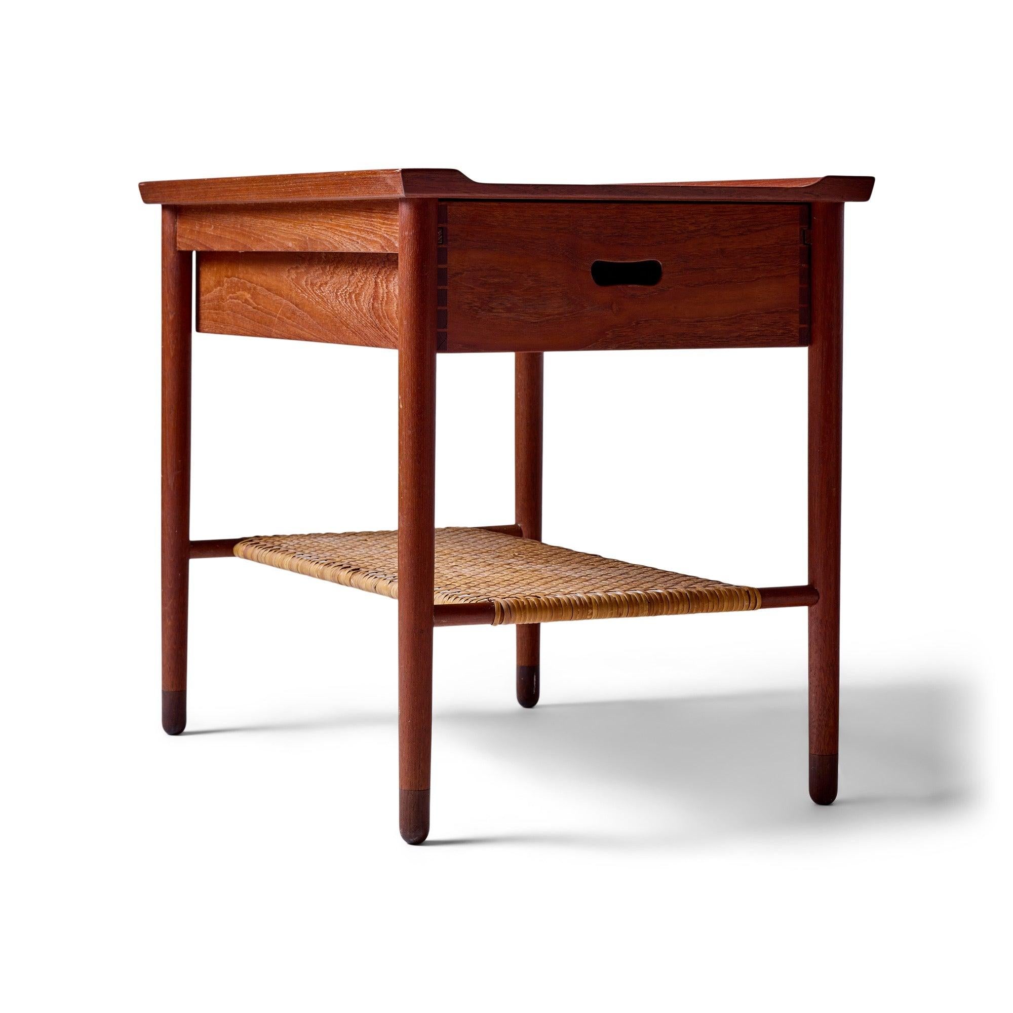 Un plateau de table à bord surélevé repose sur un tiroir coulissant à double sens, présentant une menuiserie à queue d'aronde, une poignée découpée et un plateau de rangement intérieur coulissant. Au-dessous, une étagère en rotin, soutenue par des