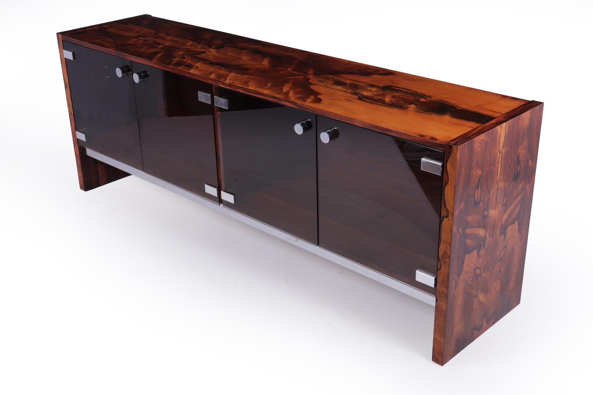 Wood sideboard by Merrow Associates c1960
Designed by Richard Young and produced by Merrow Associates having smoked glass doors with adjustable glass shelves behind. This sideboard has been produced in the finest quality Rosewood veneer with chrome