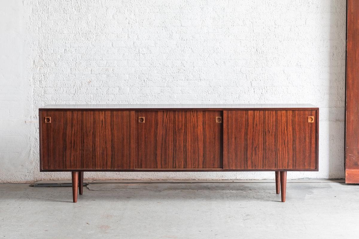 Sideboard designed and produced in Denmark around 1960. The cabinet has a rosewood veneer body with 3 sliding doors. In the middle part you can find drawers, left and right you have shelves. In good condition as shown in the pictures.

H: 76 cm
W: