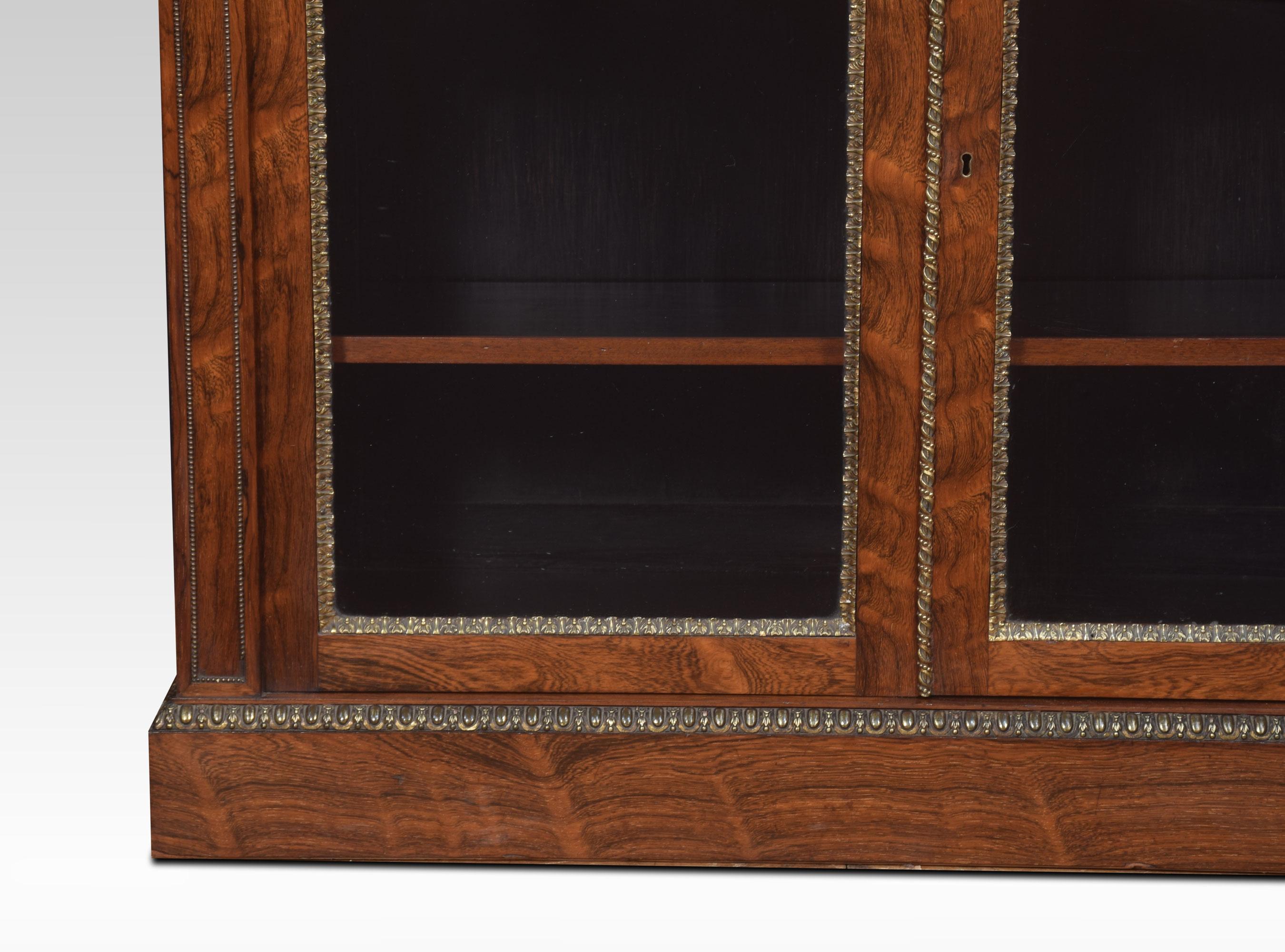 Two-door bookcase by Holland and Sons. The well-figured top having brass mounts. Above a pair of glazed doors opening to reveal an adjustable shelved interior. Flanked by molded columns with brass border. All raised up on plinth