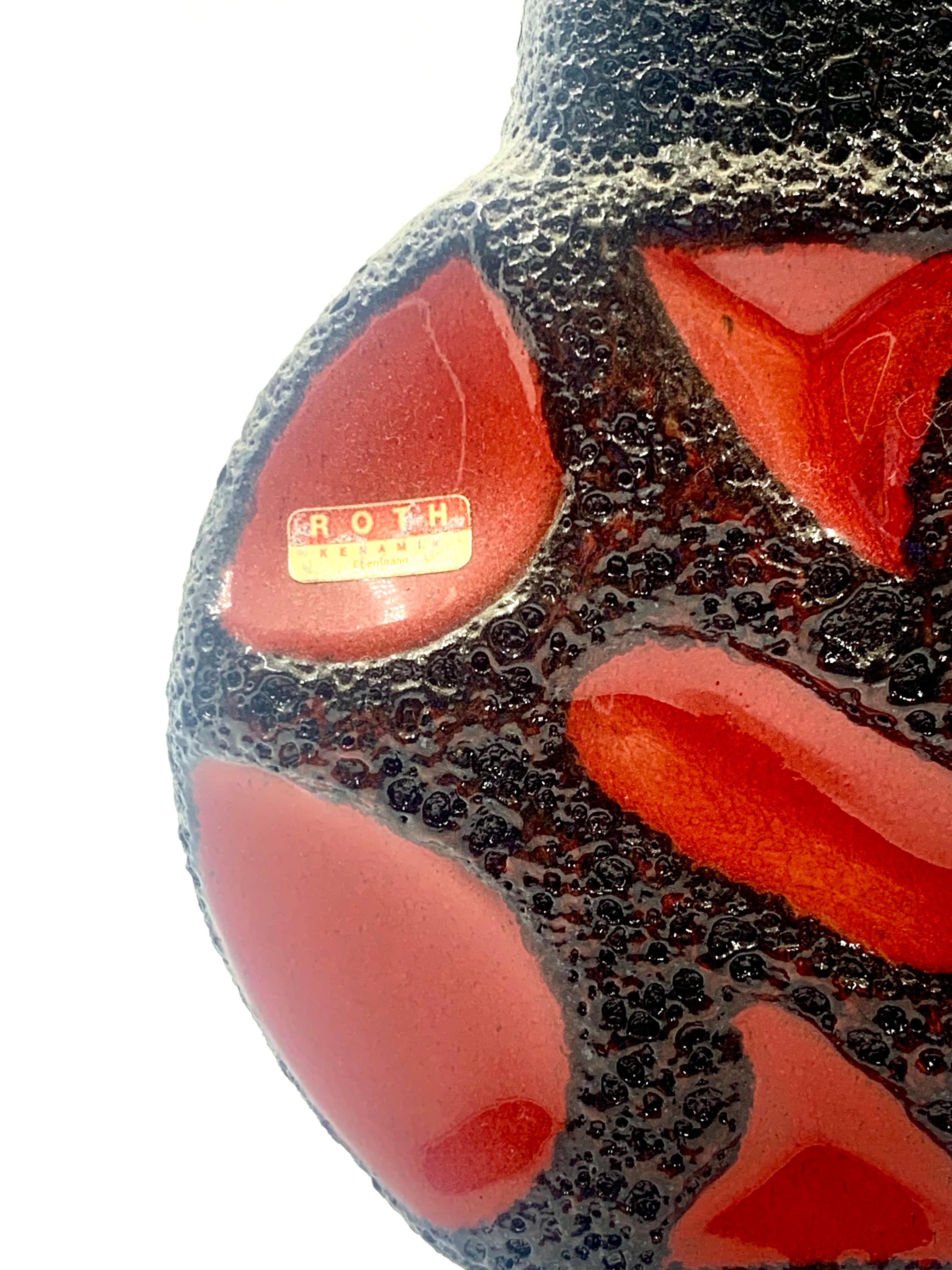 Un jarrón Roth Keramic de dos asas anulares en rojo brillante con bordes esmaltados en lava negra, número de modelo 313.
Se fabrica en dos tamaños, de los cuales éste es el más pequeño.
Conserva la etiqueta de aluminio del fabricante.