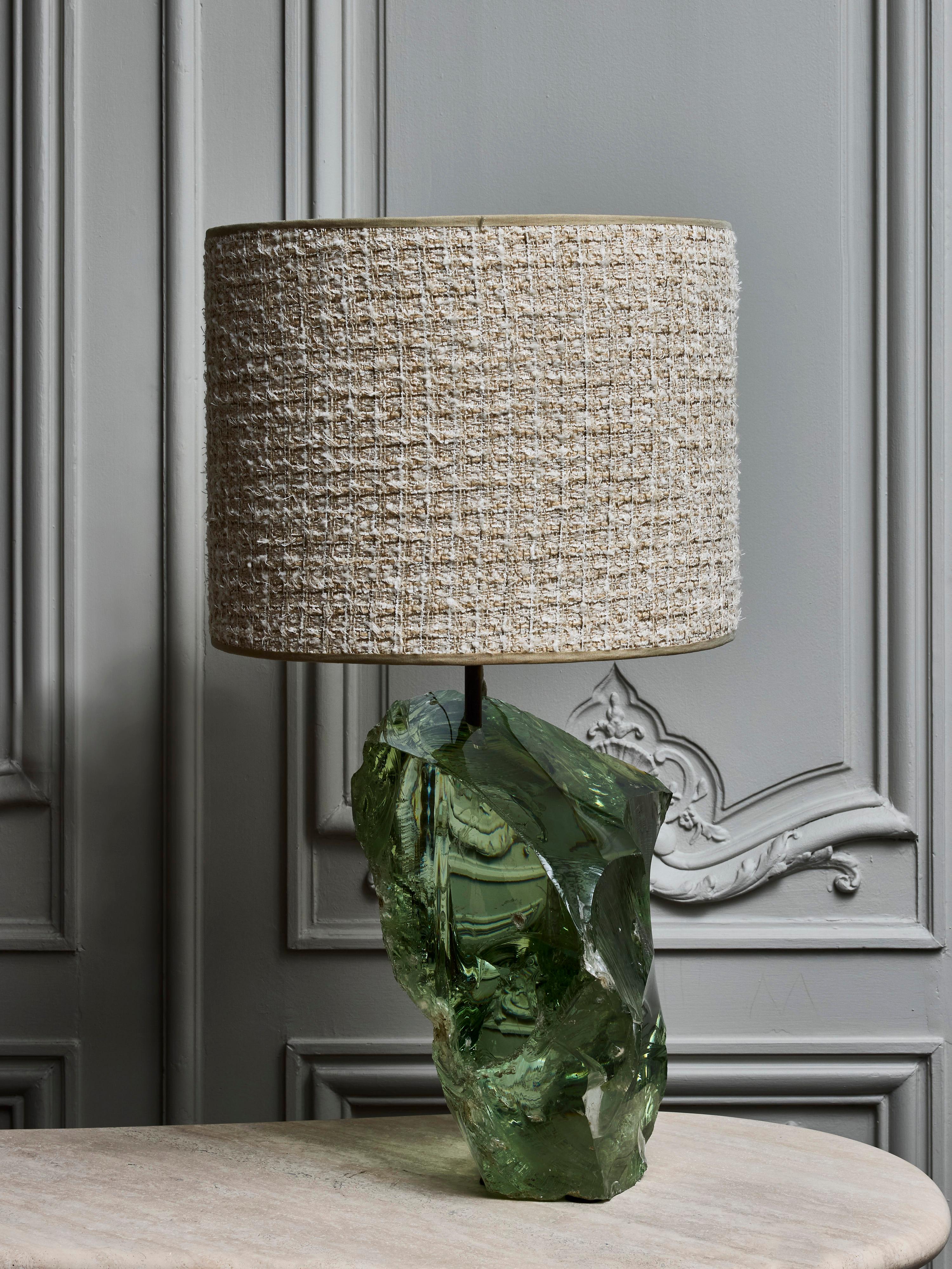 A large table lamp crafted from a substantial block of light green glass features an asymmetrical shape with rugged edges, adorned with a contemporary lampshade.