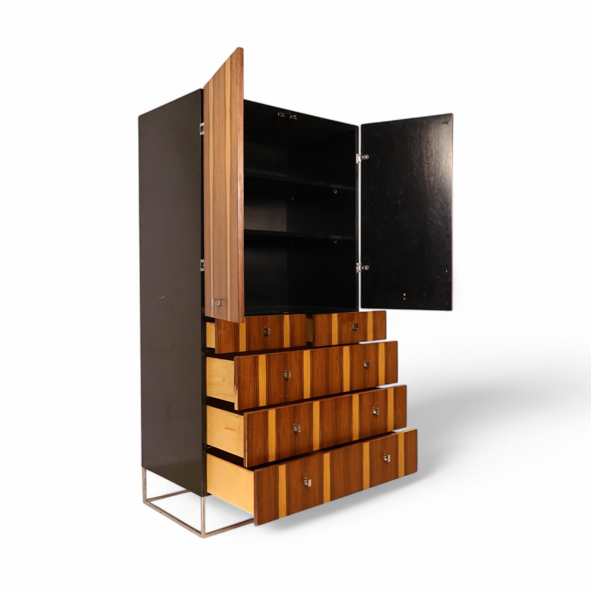 This vintage mid-century modern rosewood and chrome armoire by Rougier combines striking aesthetics with practical storage. The rich rosewood veneer, with its striking vertical grain patterns, creates a bold visual statement. Set atop a minimalist