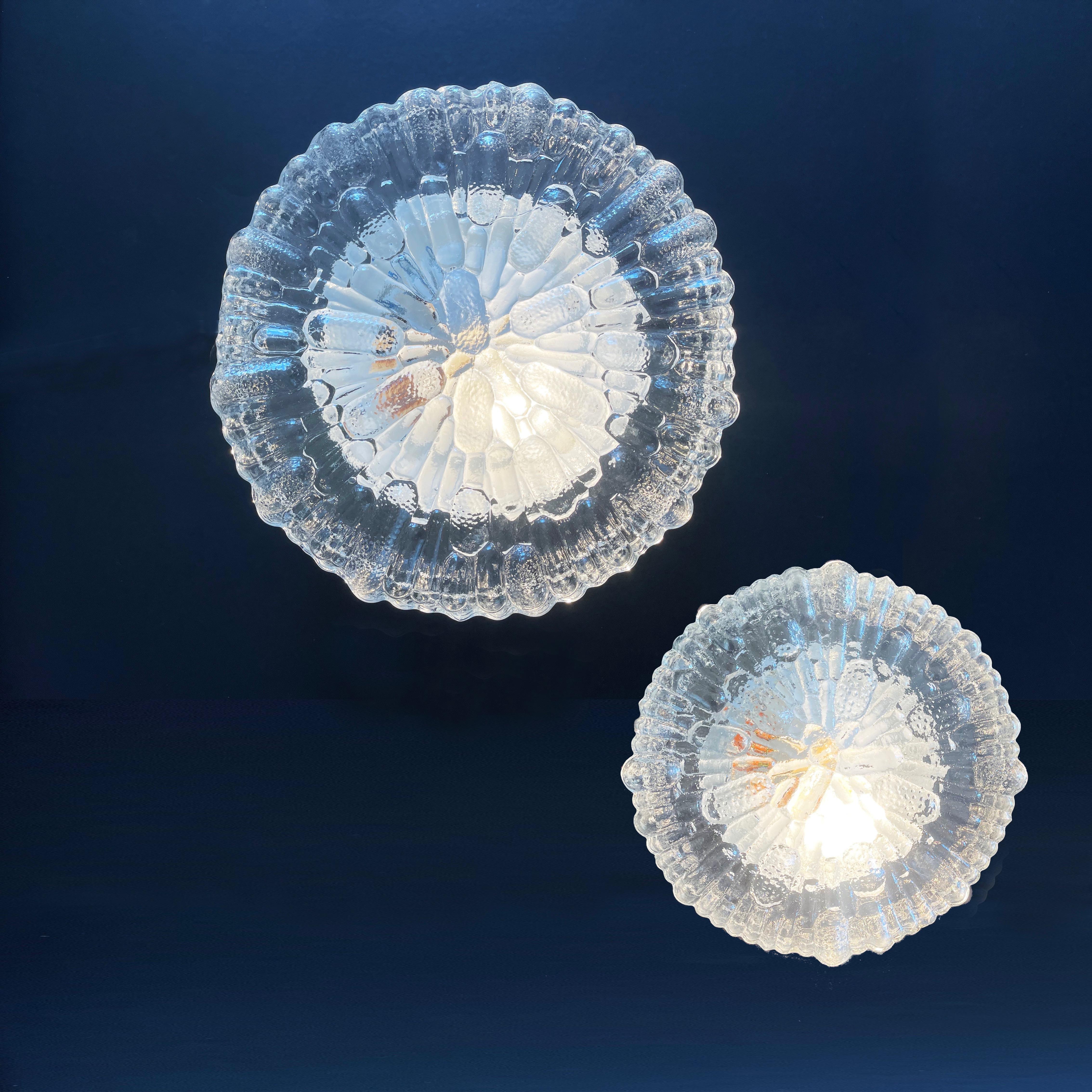 Abstract reed patterns emanating like a sunburst, a set of 2 flush base ceiling or wall lamps, made by 
Eurolux Mand in the 1970s.
They in a round, geometrical bubbly disk shape from clear glass.

The funky glas lampshades are sitting on a metal