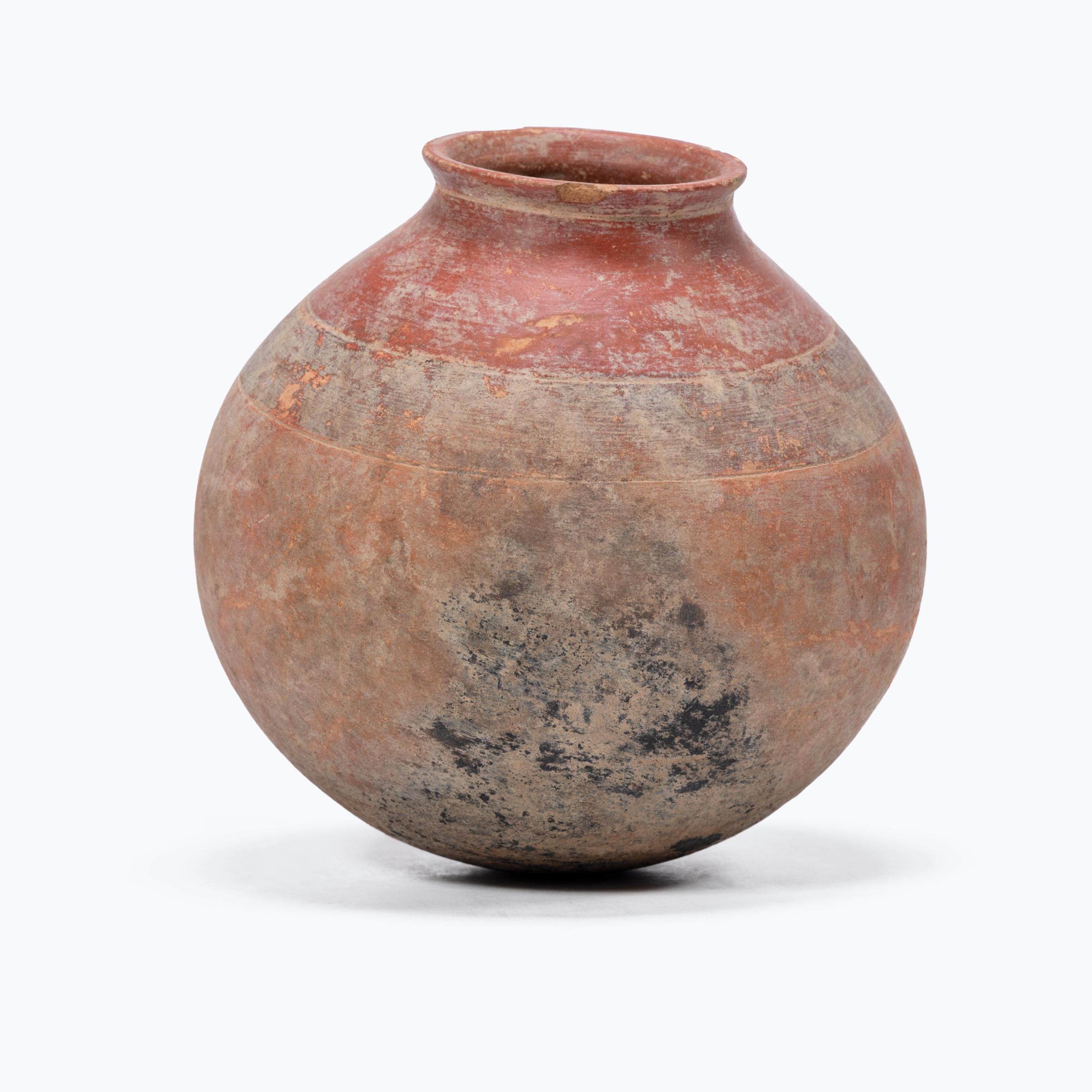 Covered in a beautifully worn red clay slip, this hand-formed ceramic pot is an excavated redware vessel from West Africa. Horizontal bands of colored slip partition the vessel's rounded form, interrupted by dark smoke residue clouding the surface.