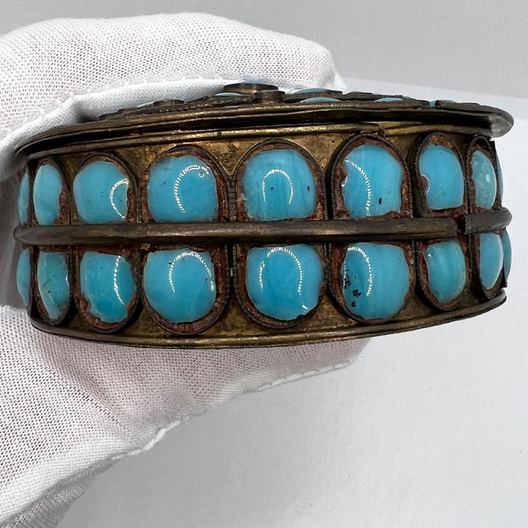A small round metal decorative box with a lid, and inlaid glass or stones in blue. This piece comes from Tibet and is created from brass. The top and sides are decorated with polished glass or blue semi-precious stones. The lid is unhinged and opens