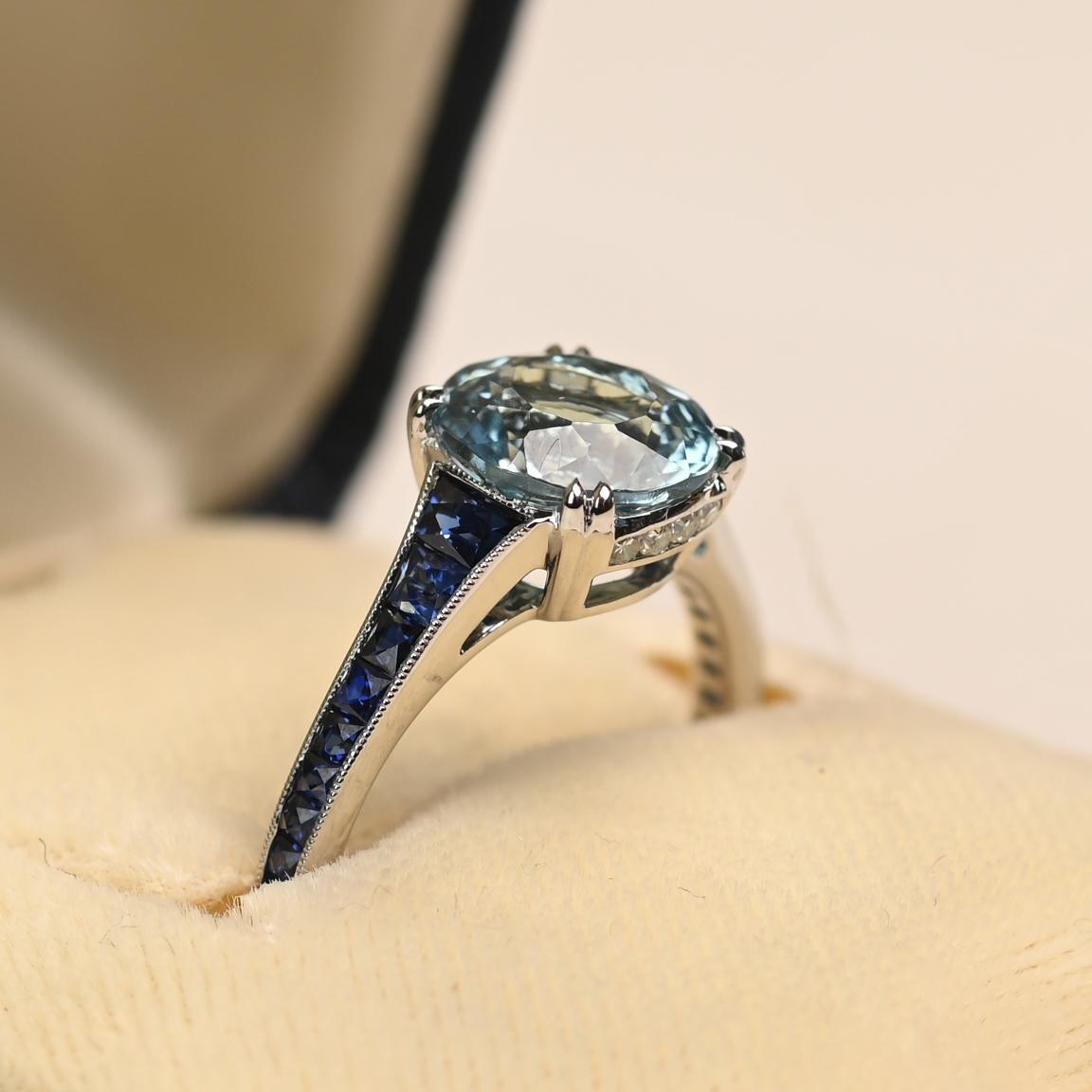 Cette élégante bague met en valeur une vibrante aigue-marine ronde pesant environ 1,70 carat, fixée dans une monture raffinée à deux griffes. Les épaules sont ornées de saphirs de taille française, totalisant environ 0,60 carats, rehaussés de