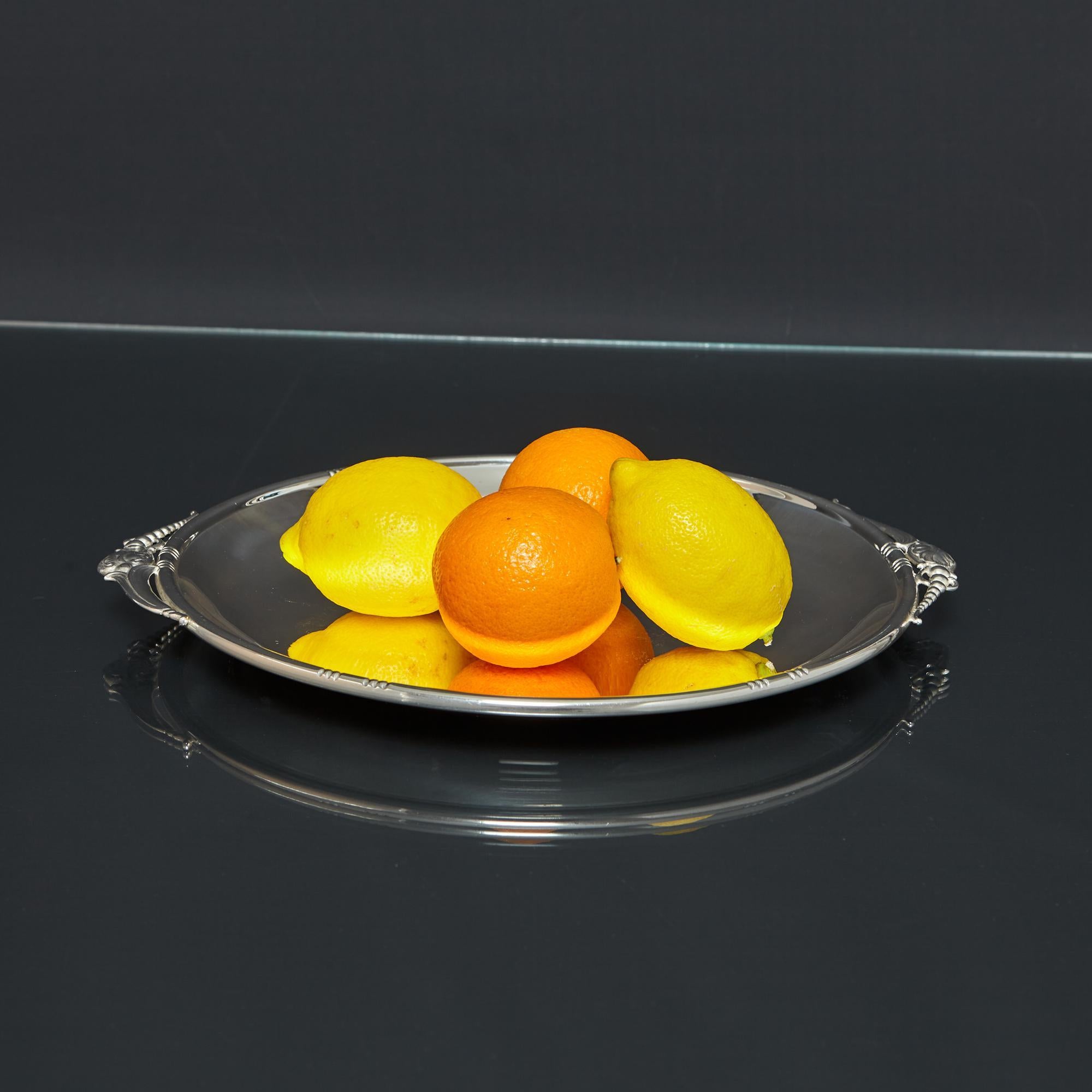 Plato o bandeja redonda y poco profunda, de plata de ley, de estilo Art Nouveau. La sencillez del estilo se realza con asas caladas de fundición, cuentas graduadas aplicadas, volutas y hojas, todo ello evocador de la época Art Nouveau.

Con