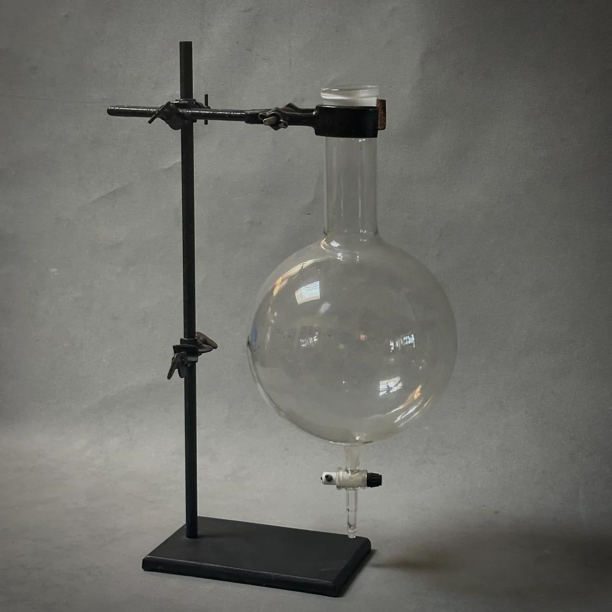 This early 20th-century Belgian scientific apparatus, featuring a tear-drop glass beaker set upon a stark metal stand, showcases an elegant fusion of form and function. The round beaker, with its delicate, bubble-like quality, contrasts starkly