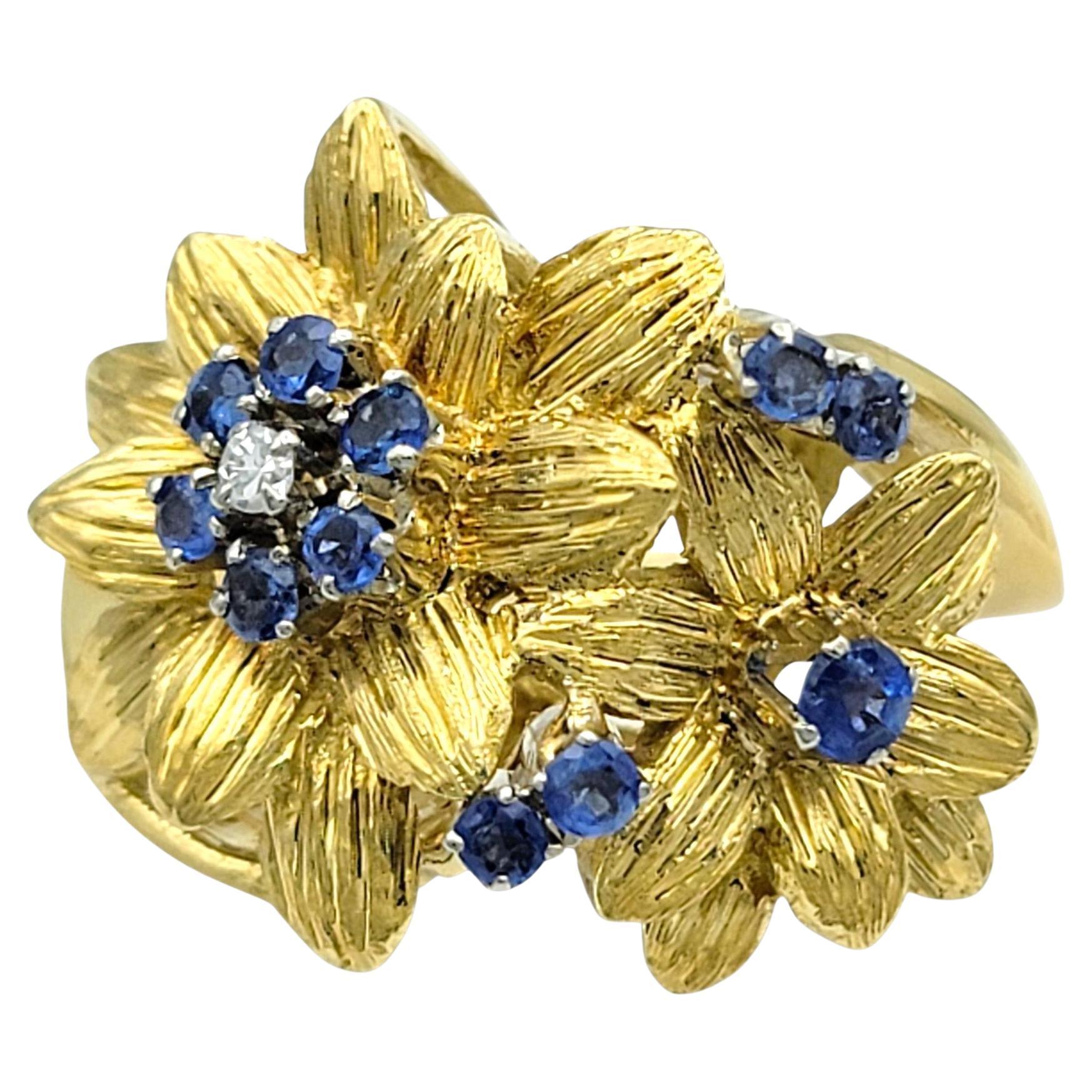 Cette charmante bague présente un motif floral détaillé avec des pétales d'or texturés qui créent un aspect doux et dimensionnel. Au centre de chaque fleur se trouve un petit éclat de saphirs d'un bleu profond et de diamants blancs, ajoutant un