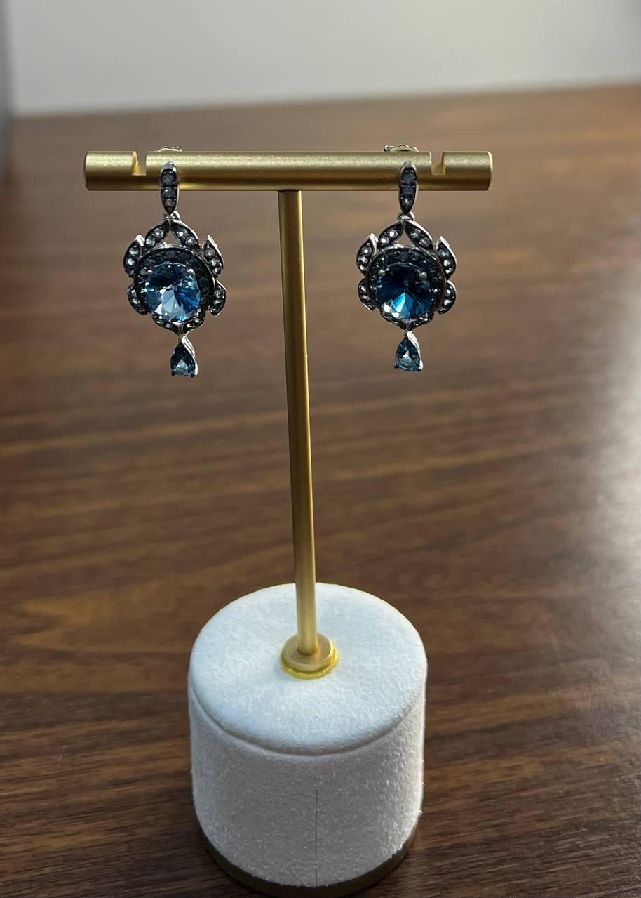 These drop earrings feature round blue stones set in 14k White Gold. The design includes intricate detailing around the stones, with additional small diamonds accenting the piece.
