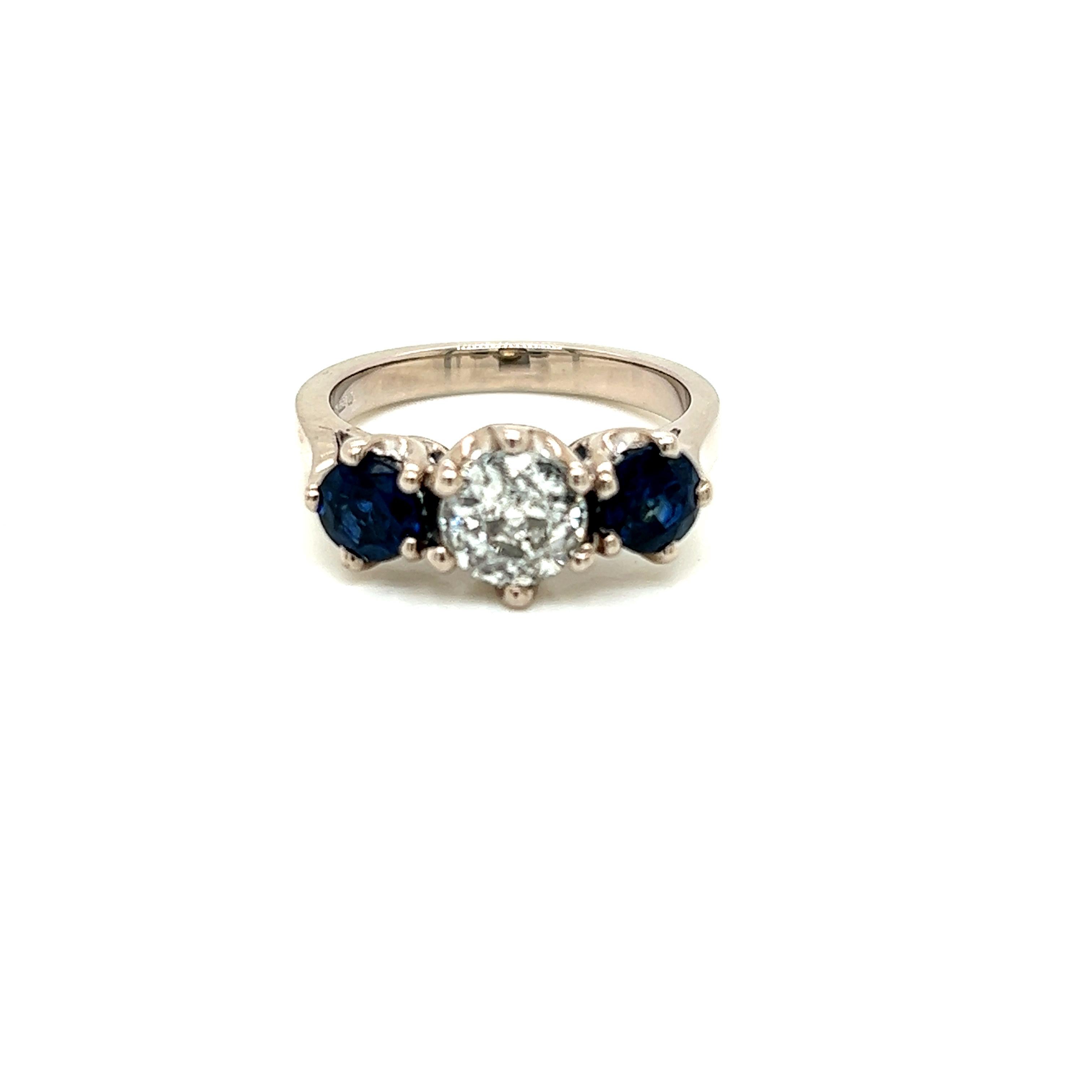 This charming ring features a lovely 0.85 carat Round Brilliant Diamond with a Round Brilliant Blue Sapphire on either side of it set on an 18K White Gold band.

The diamond at the centre of the ring weighs 0.85 carats and is of J colour and I1