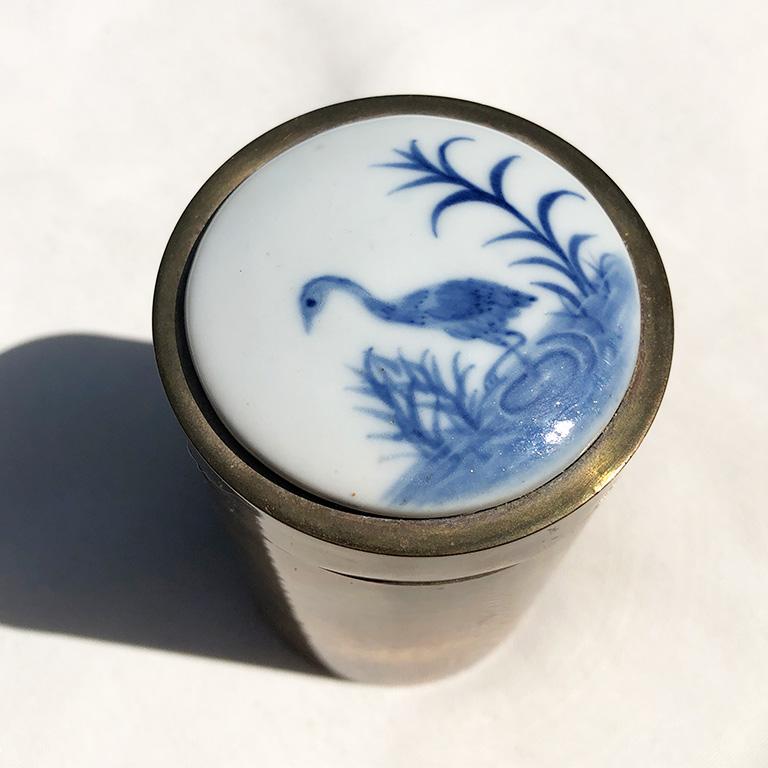 Round ceramic and brass container with a brass bottom and removable lid with ceramic top. Top features a blue and white ceramic or porcelain lid with floral hand-painted and glazed depiction of a crane amidst floral detail background. A beautiful