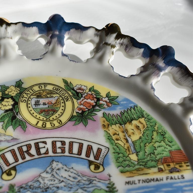 Unique ceramic circular blue and gold souvenir plate from Oregon. This beautiful plate is round in form, and features scalloped pierced edges. The piercings are quatrefoil in shape. The edges almost resemble a starburst in how they extend from the