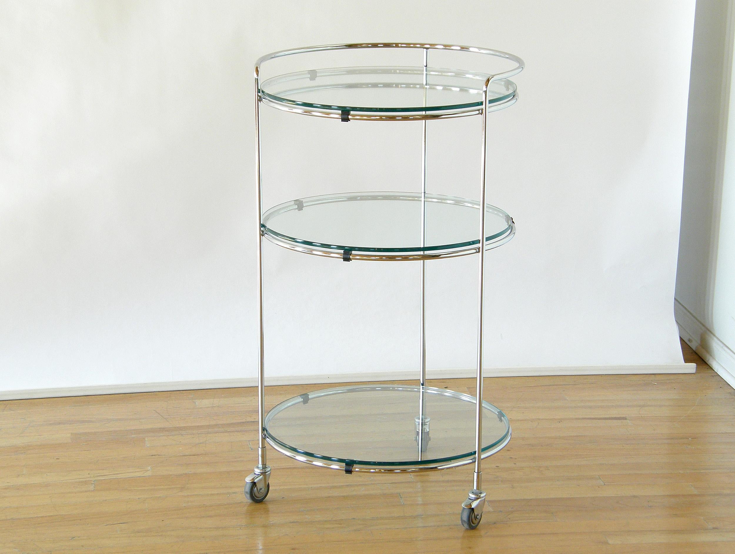 This compact, chrome serving cart has an elegant, Minimalist style. The slender, tubular chrome forms a skeletal framework that supports three glass shelves. The chrome extends above the top shelf part way around. It bends to form a rim that can be
