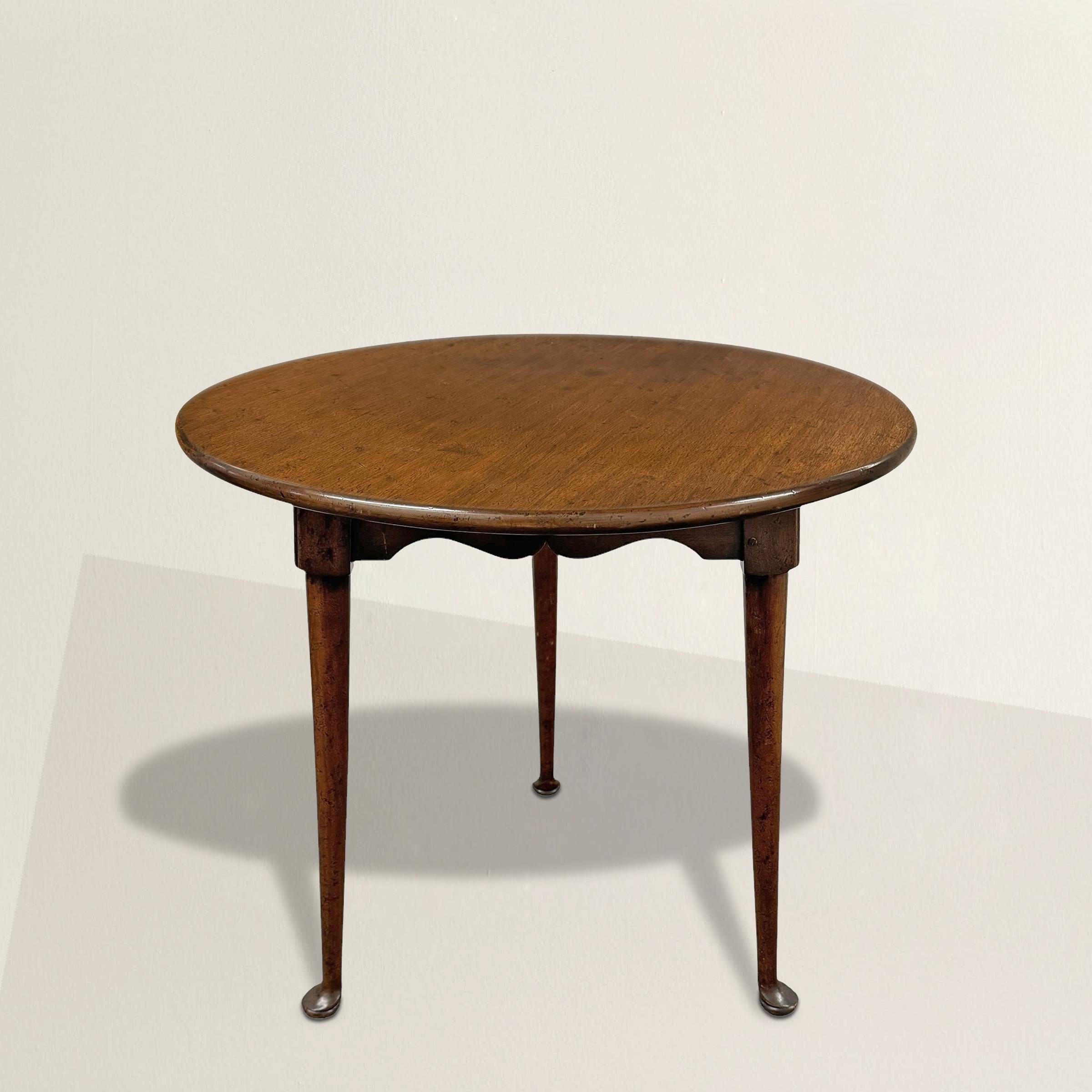 This charming 20th-century American walnut cricket table is a loving nod to 18th-century Queen Anne design. With its round top, scalloped apron, and three elegantly tapered legs ending in classic pad feet, it captures all the grace and simplicity of