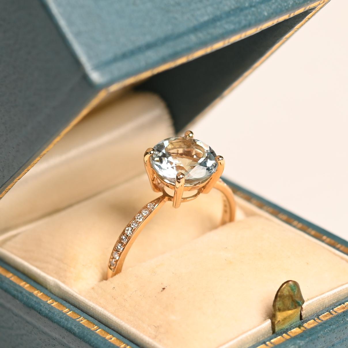 Este exquisito anillo de compromiso exhibe una impresionante aguamarina talla redonda de aproximadamente 1,9 quilates, elegantemente sujeta en garras. El delicado tono azul de la gema irradia una belleza atemporal, convirtiéndola en una cautivadora