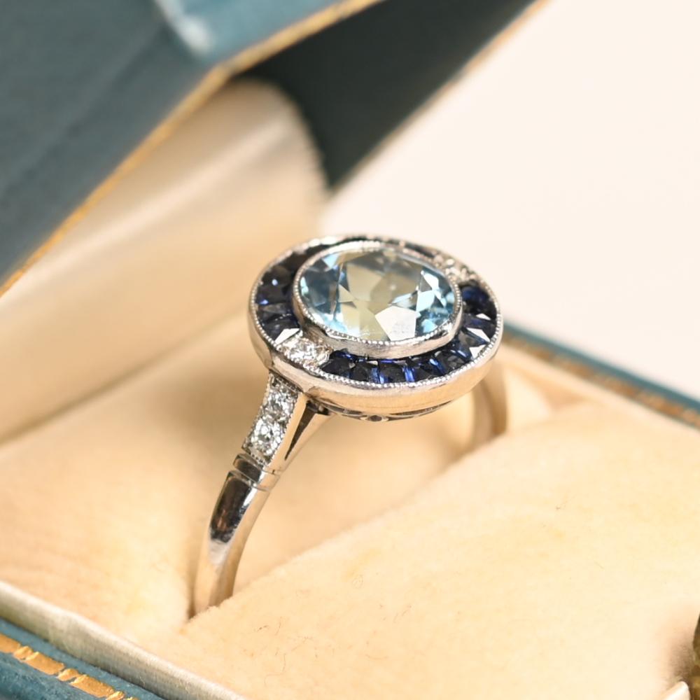 Cette bague exquise est ornée d'une superbe aigue-marine de 1 carat environ, de taille ronde, qui rayonne d'une teinte bleue sereine. L'aigue-marine est entourée d'un étonnant halo de saphirs de taille française méticuleusement disposés, totalisant
