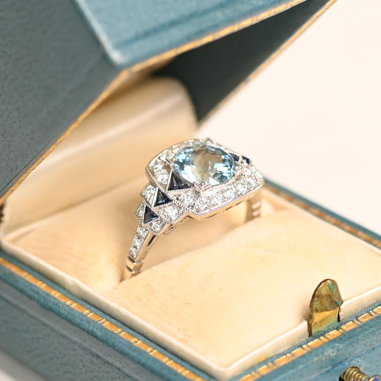 Cette exquise bague géométrique met en valeur une superbe aigue-marine de taille ronde d'environ 1,20 carat, solidement sertie dans de délicates pinces. La teinte bleue sereine de l'aigue-marine est magnifiquement complétée par une rangée de saphirs