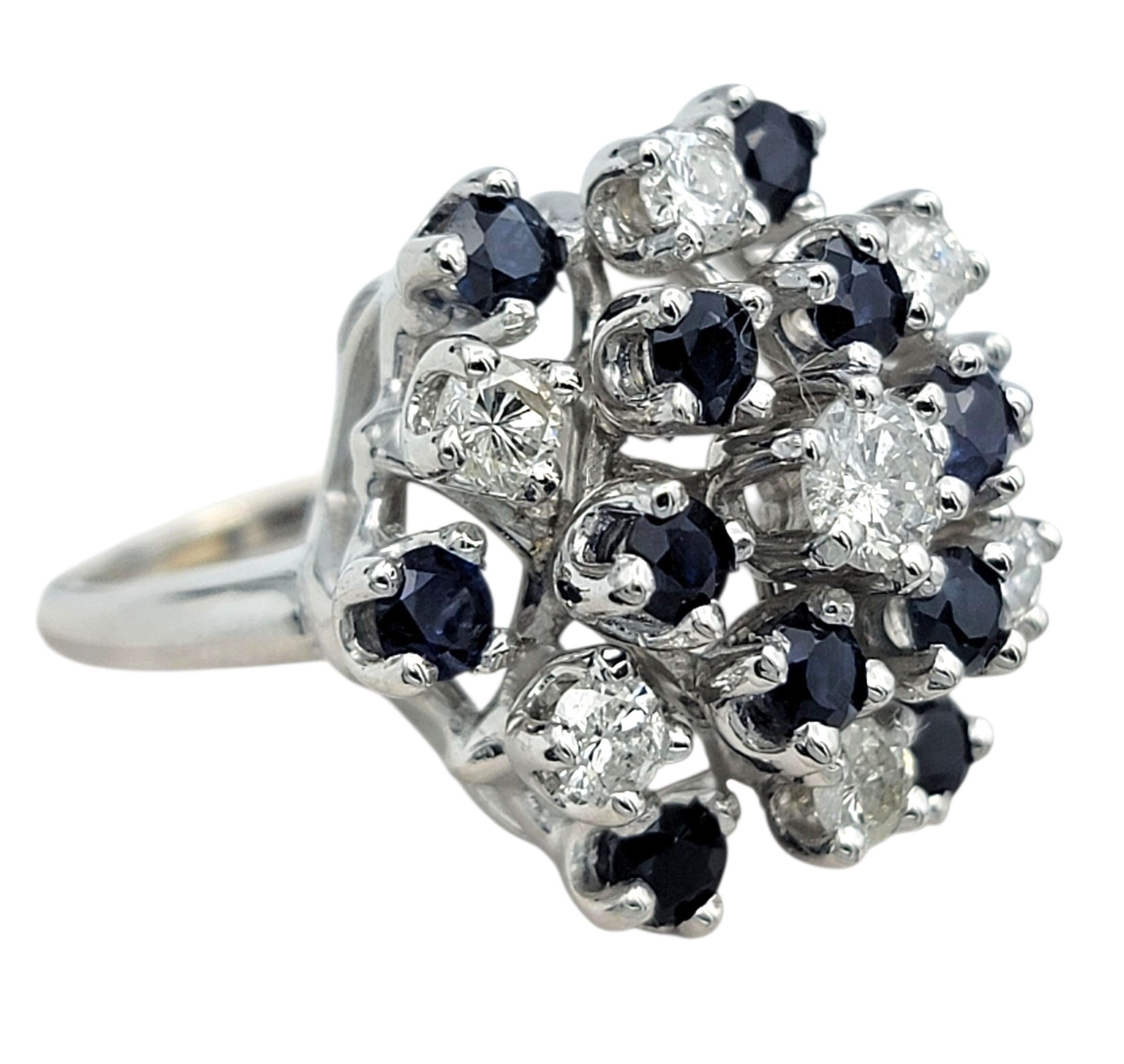 Cette magnifique bague présente un motif de grappe arrondi rempli de pierres blanches brillantes qui créent une surface douce et étincelante. Parmi eux, des saphirs bleus vifs ajoutent une couleur riche et donnent à la pièce un contraste vif et