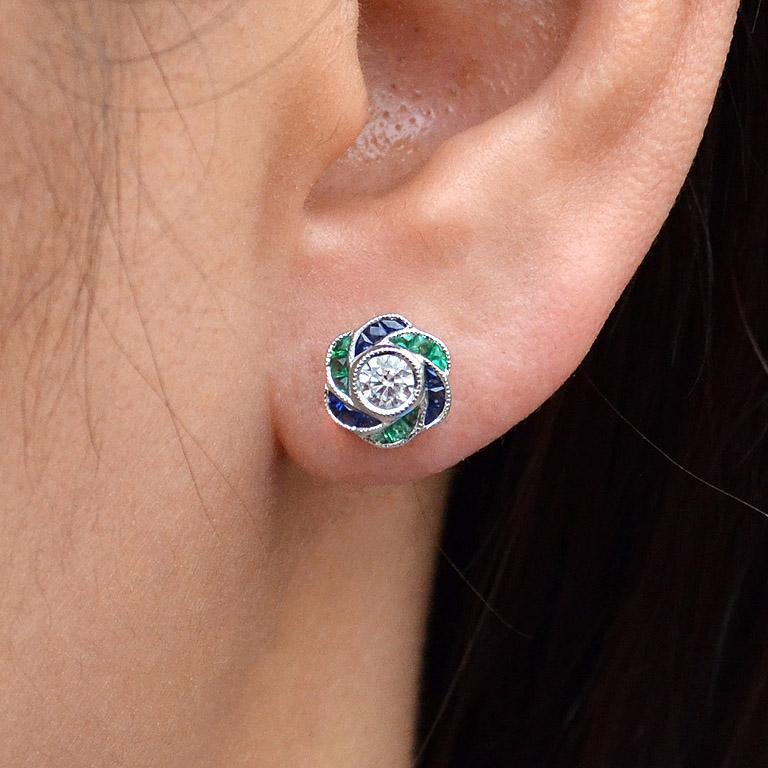Perfect with everyday wear, these charming vintage Art Deco revivalist design stud earrings feature a pair of brilliant cut diamonds surrounded by bright blue sapphire and emerald for rose petals finished look all in 18K white