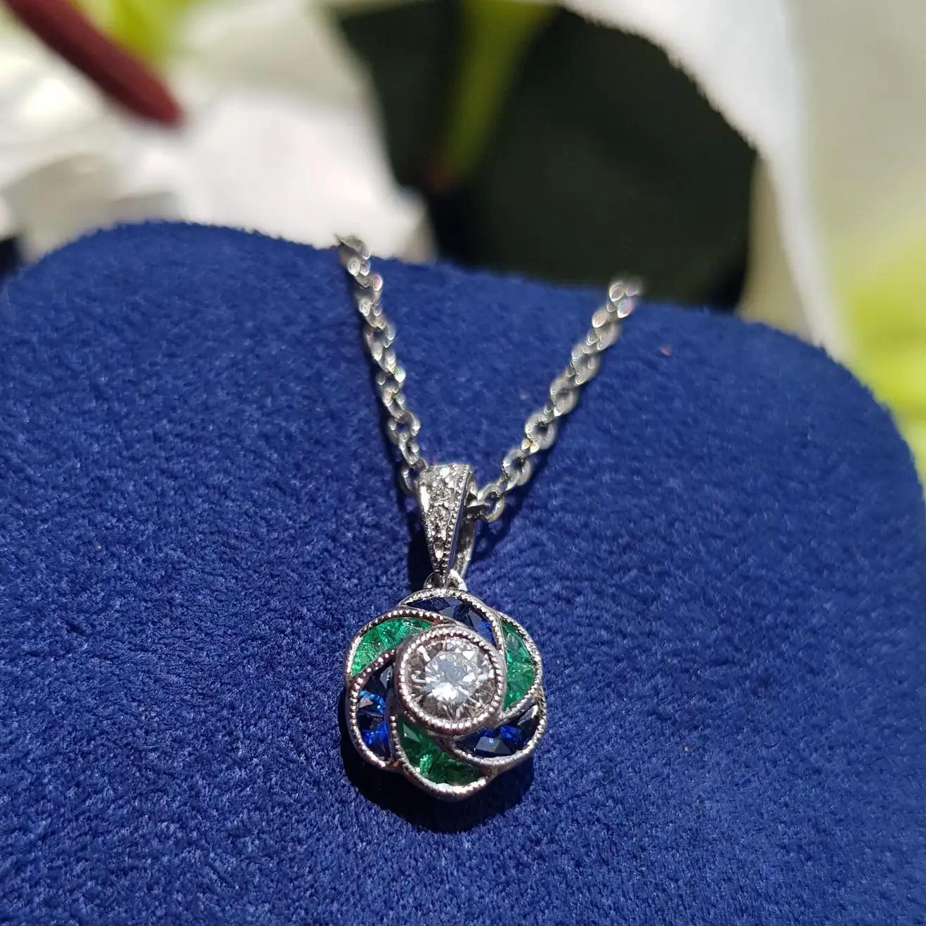 A stunning round cut diamond at the center, surrounded by alternating emerald and sapphire stones arranged in a circular pattern. The design beautifully mimics a rose flower, with the vibrant colors of the emeralds and sapphires enhancing the