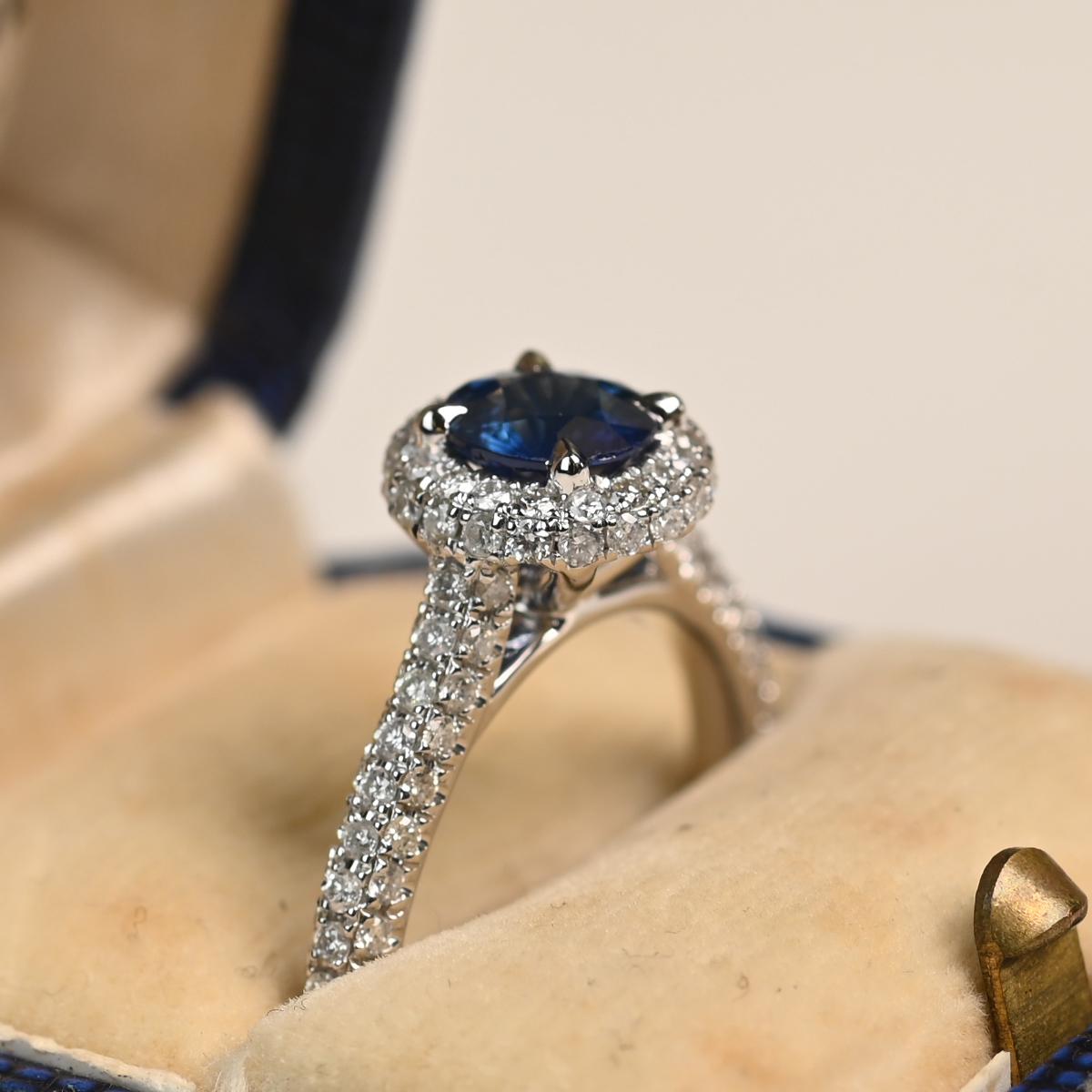 Cette bague exquise met en valeur un saphir rond vibrant pesant environ 0,90 carats, solidement serti dans une monture à griffes classique. Le saphir est entouré d'un halo bombé composé de deux rangées de diamants ronds étincelants, créant ainsi un