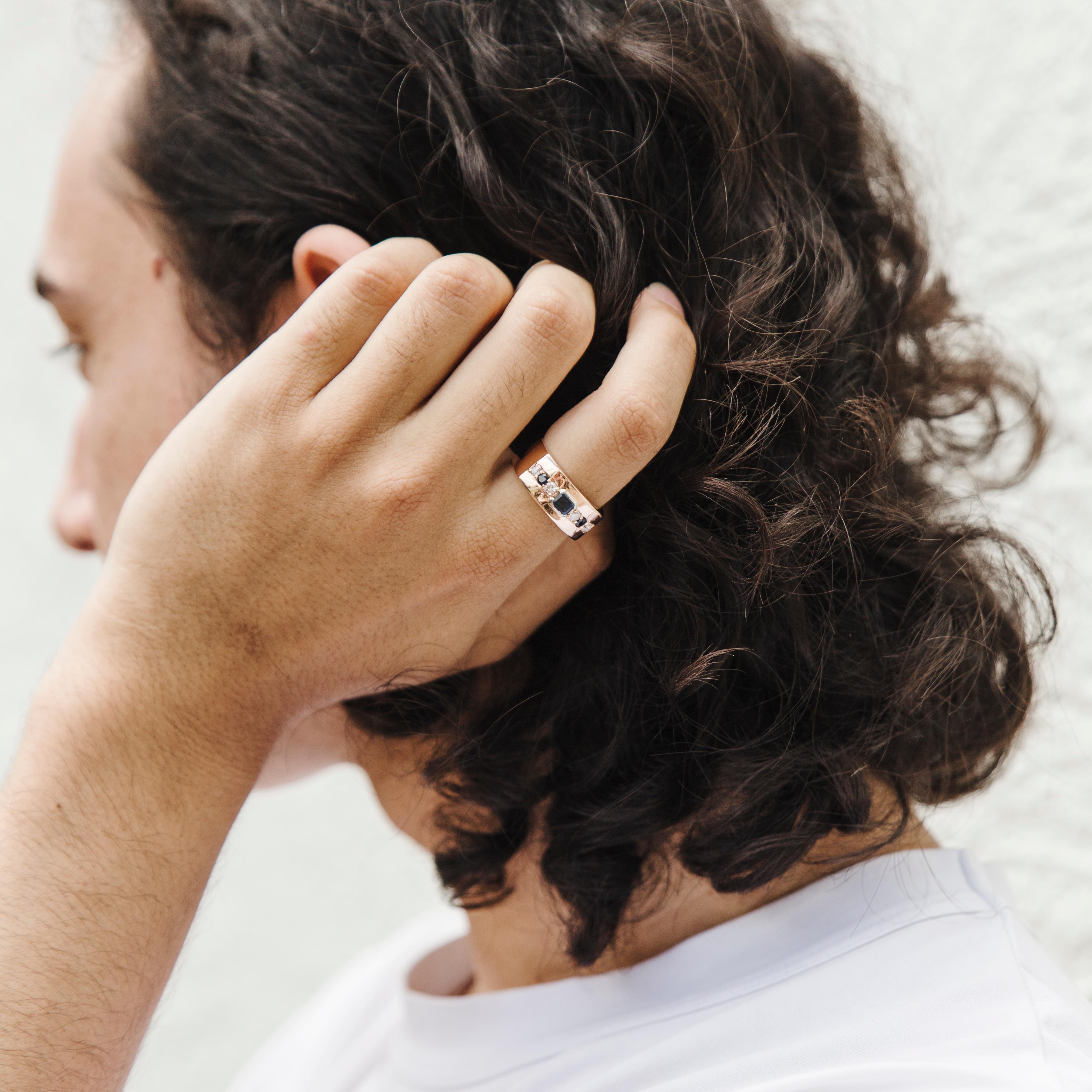 Forged in 9 carat rose gold, this handsome vintage ring sports a smooth reflective finish and set with a deep blue emerald cut sapphire flanked by two smaller sapphires and round brilliant diamonds. We have named this dapper band The Amos Ring. The