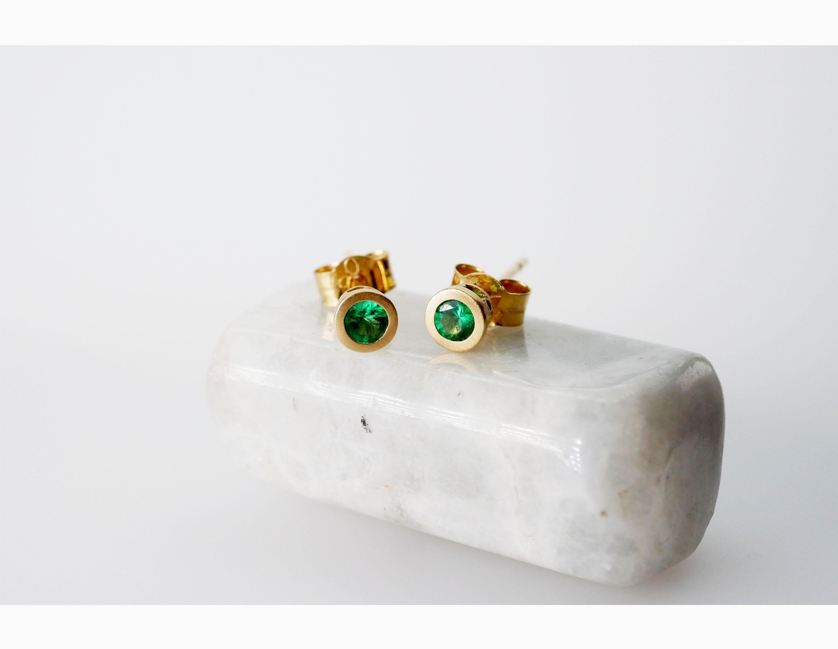Delicate bright green round emeralds stud earrings in 18k yellow gold. These earrings are set in a smooth bezel setting with a matt finish and decorative pattern that lets the light shine through the emeralds. Wear solo or stacked with other colour