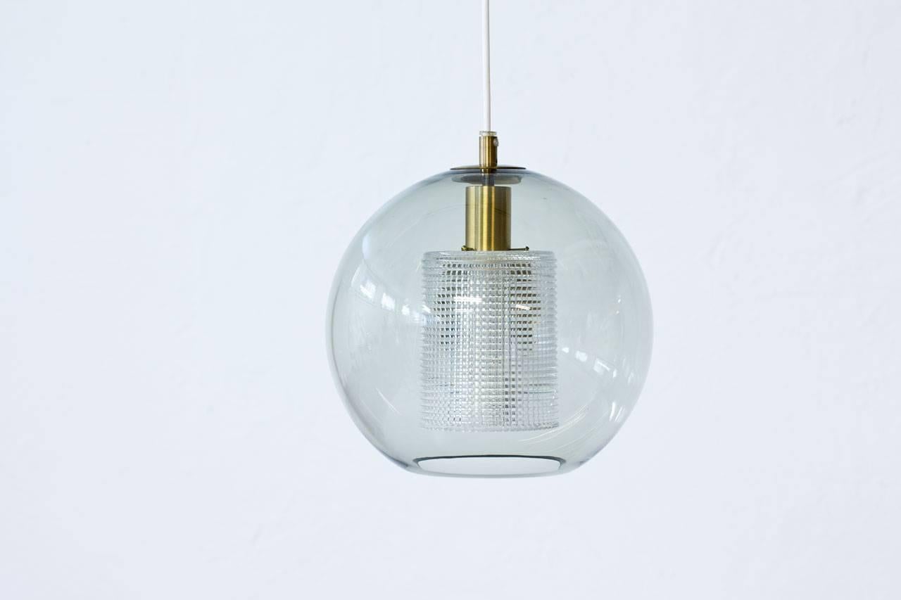 Pendant lamp designed by Carl Fagerlund for Swedish glass company Orrefors during the 1960s. Sphere shaped outer cup in clear grey tinted glass with a cylinder internal diffuser in clear pressed glass. Brass fixture.