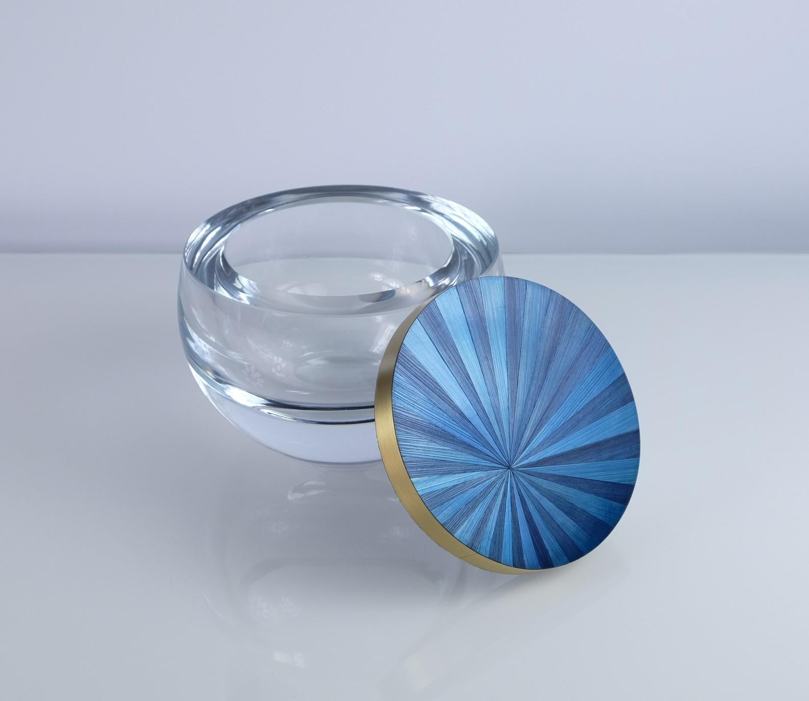This round glass box has a lid covered of a nice blue straw marquetry, surrounded with brushed brass trims. 
The radial design of the straw reminds the great Art deco period while the very chic blue color contrasts perfectly with the gold brass