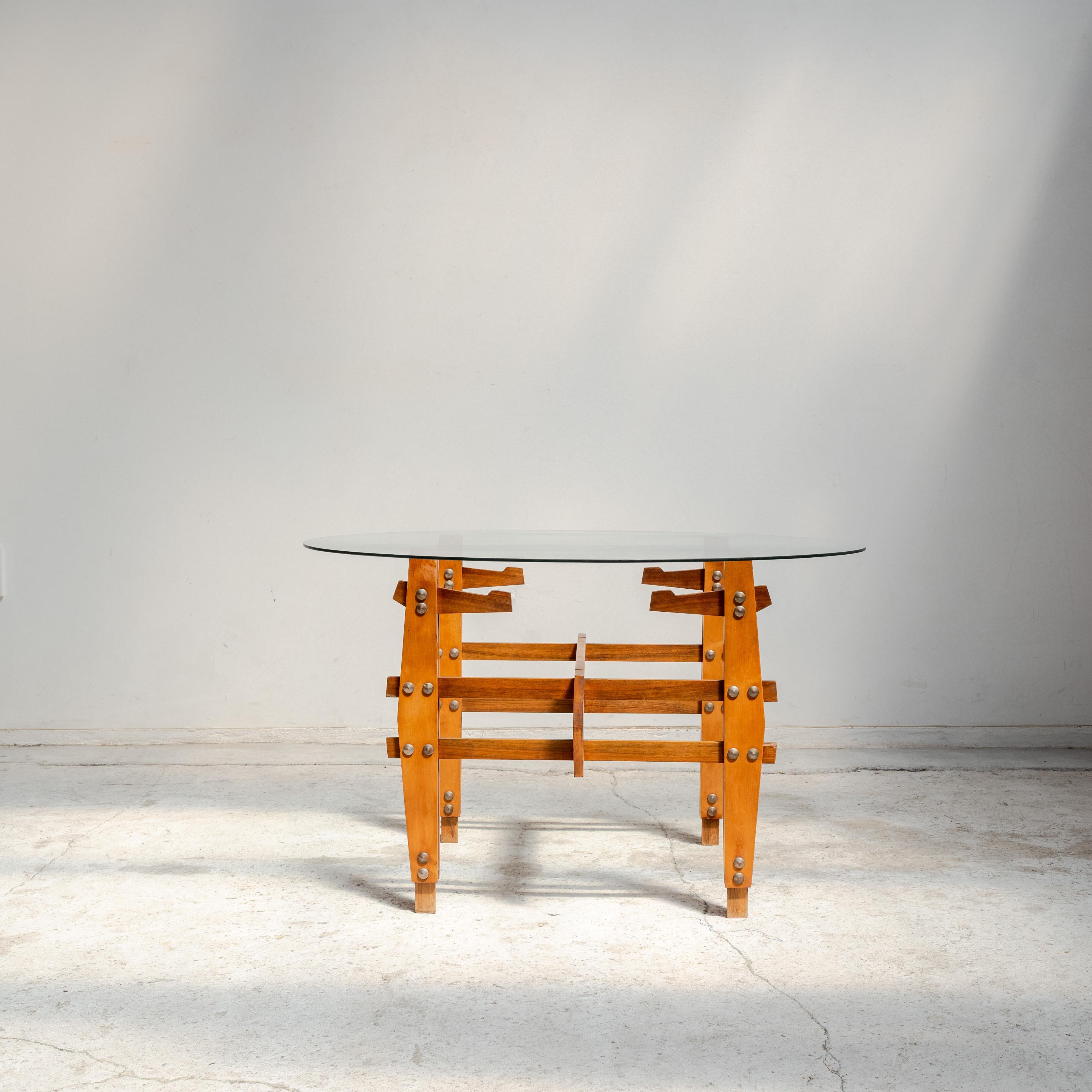 Cette table basse ronde à plateau en verre illustre l'élégance du design italien du milieu du siècle. La base sculpturale en bois, construite à partir de poutres emboîtées avec des boulons métalliques apparents, supporte un plateau circulaire en