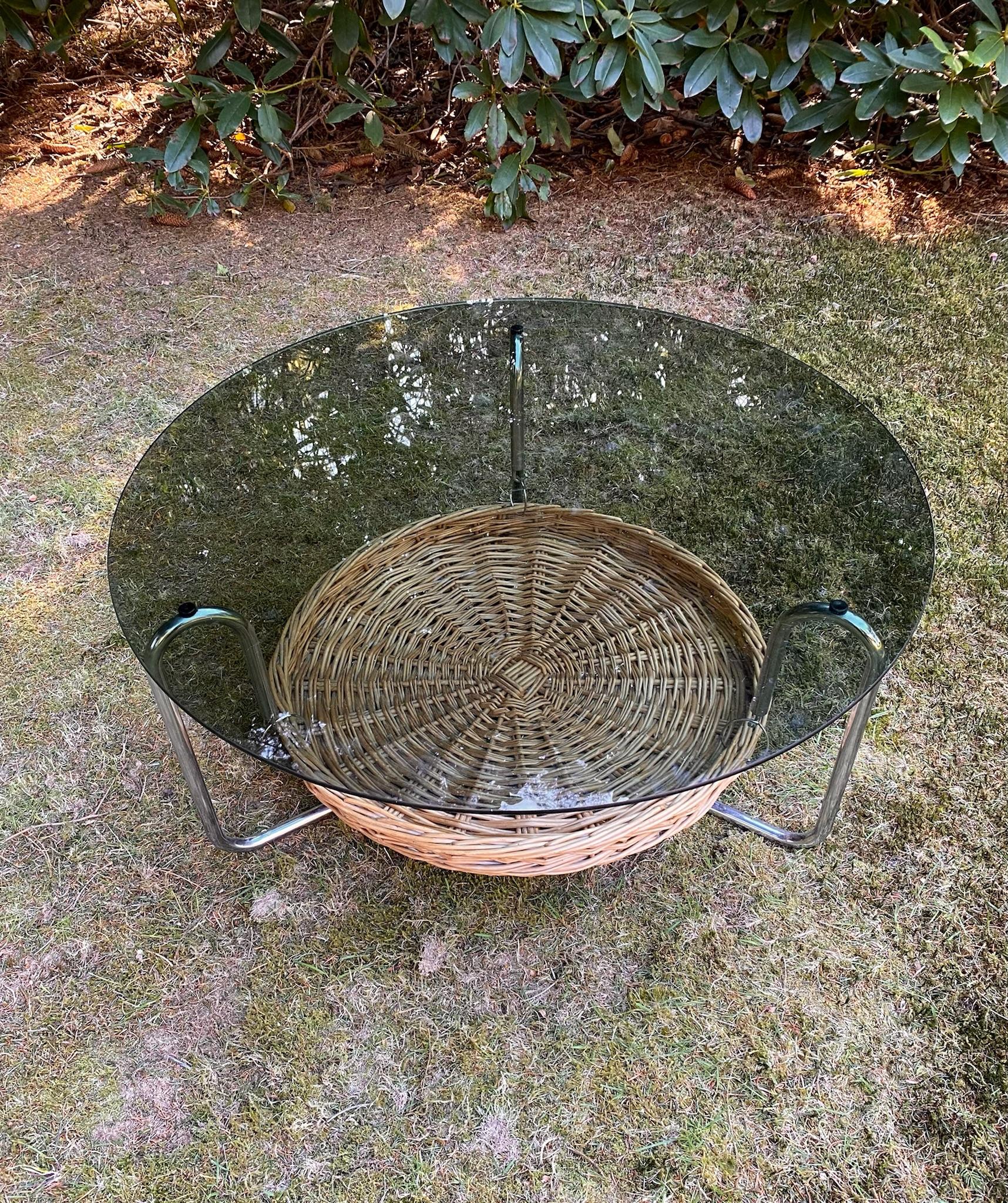 Table basse en verre avec porte-revues datant d'environ 1960. Cette table basse vintage dispose d'un magnifique cadre en métal chromé et d'un plateau en verre fumé. En dessous se trouve un panier de magazines en osier. Cette table basse est en bon