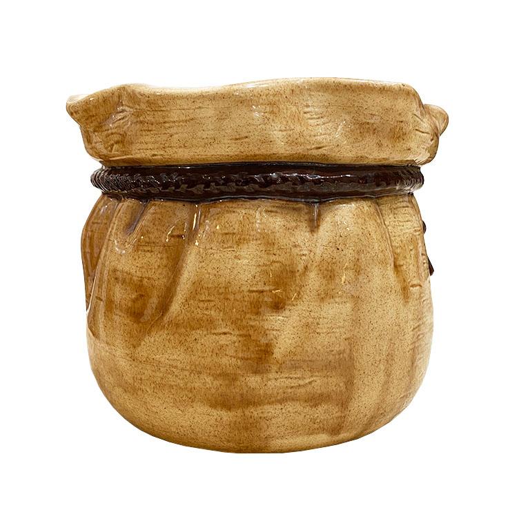 A brown ceramic planter with a scalloped edge at the top. A ceramic ribbon is sculpted around the edges with tassels in a knot that hang down. The brown on the body of the planter is a lighter brown than the tassel tied and cinched around the top,