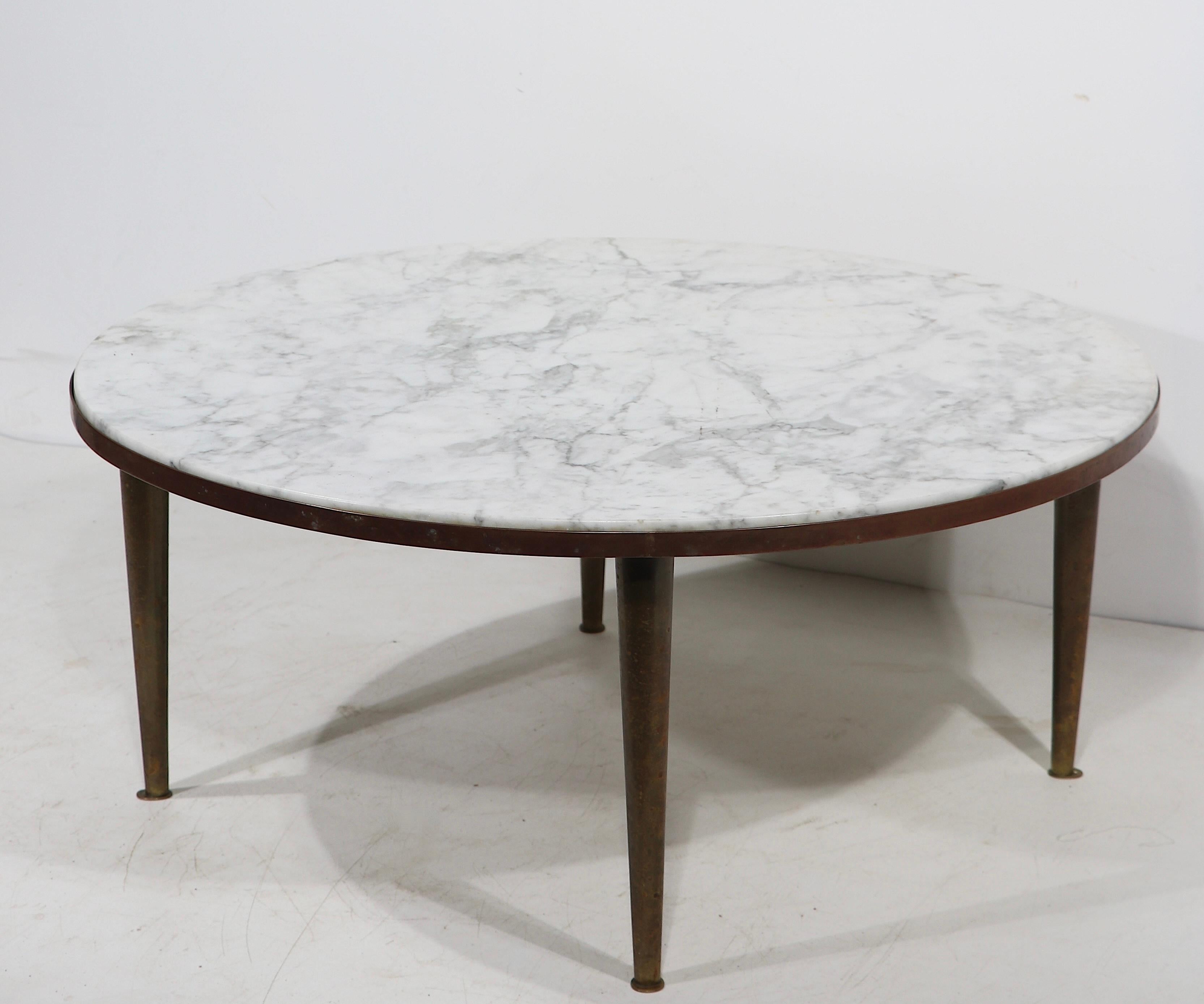 Voguish, chic, and sophisticated Mid Century coffee table featuring a thick round marble top, which rests in a brass frame on tapering brass pole legs, with unusual brass pad form feet. The table is solid, sturdy, clean and ready to use, it shows