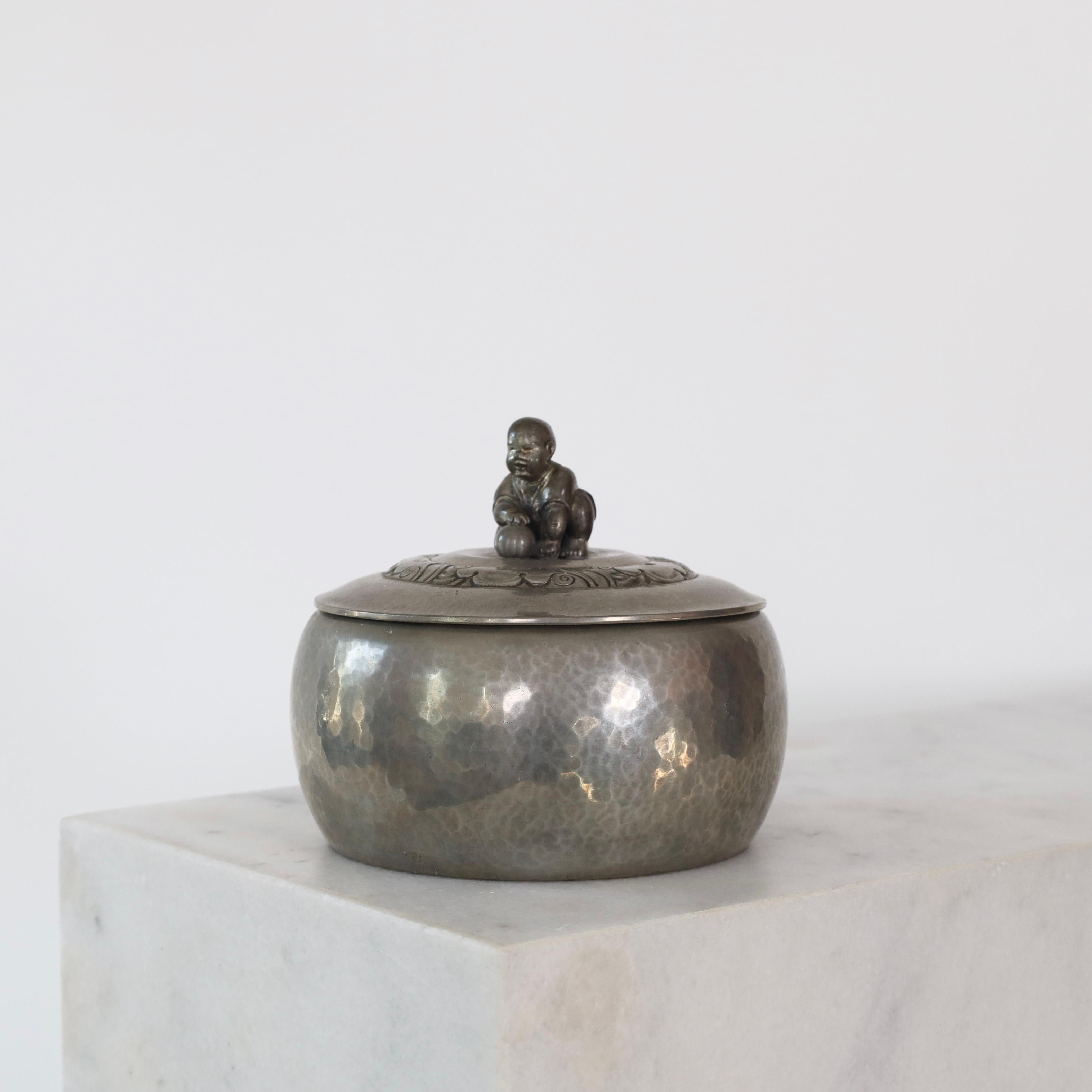 A lidded Pewter Box made by Just Andersen in the 1920s. A lovely piece for a beautiful spot.

* A round lidded box in hammered pewter with inner box in wood. The lid has ornaments and a handle shaped as a small boy with a ball.
* Designer: Just