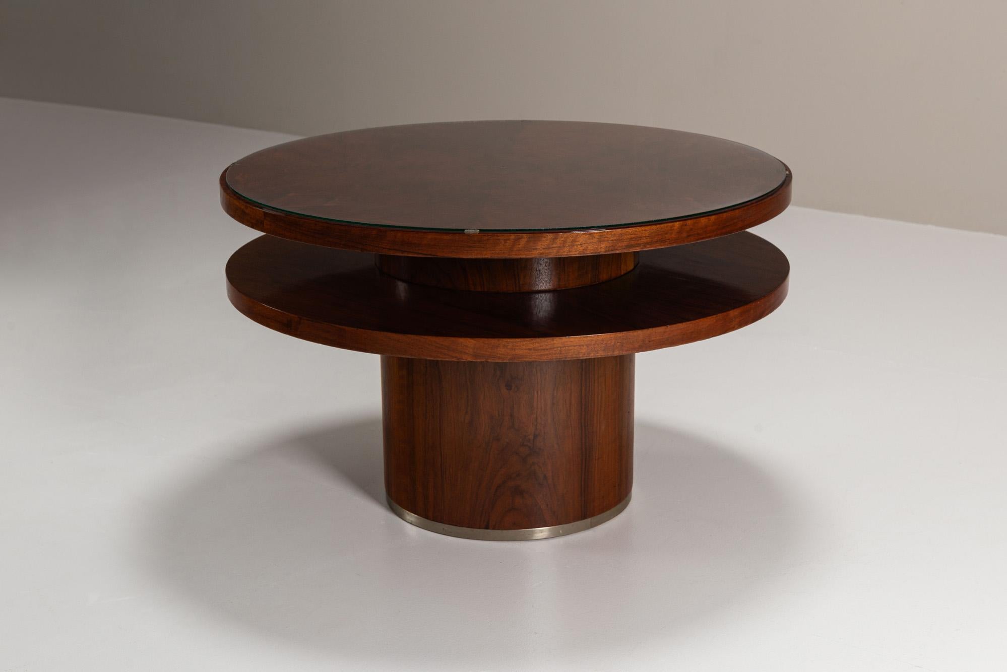 This circular table is veneered in richly figured mahogany, its top displaying an intricate play of grain beneath a protective glass plate. The glass not only safeguards the surface but also highlights the depth and character of the veneer. A second